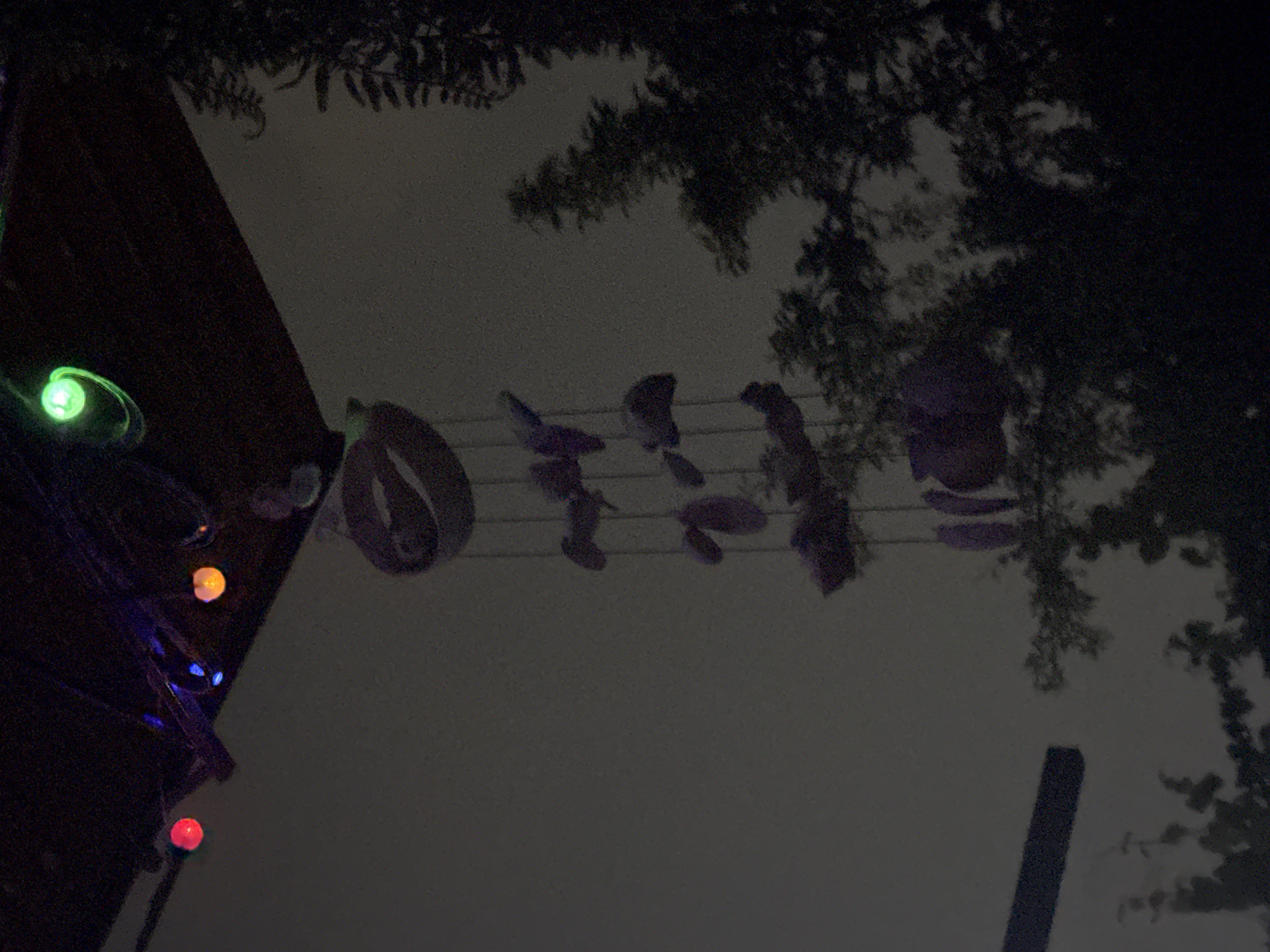 The image depicts an outdoor scene at night, possibly in Vostok, Kyrgyzstan. In the foreground, a wind chime made of purple, heart-shaped ornaments hangs from a dark structure overhead. The structure is adorned with colorful lights, including red, orange, blue, and green bulbs, some of which are blurred due to motion or low light. In the background, silhouetted trees are visible against a dimly lit sky. A dark, rectangular object, possibly a beam or a sign, is mounted horizontally on the left side of the frame. The overall impression is one of a decorative outdoor setting at night.