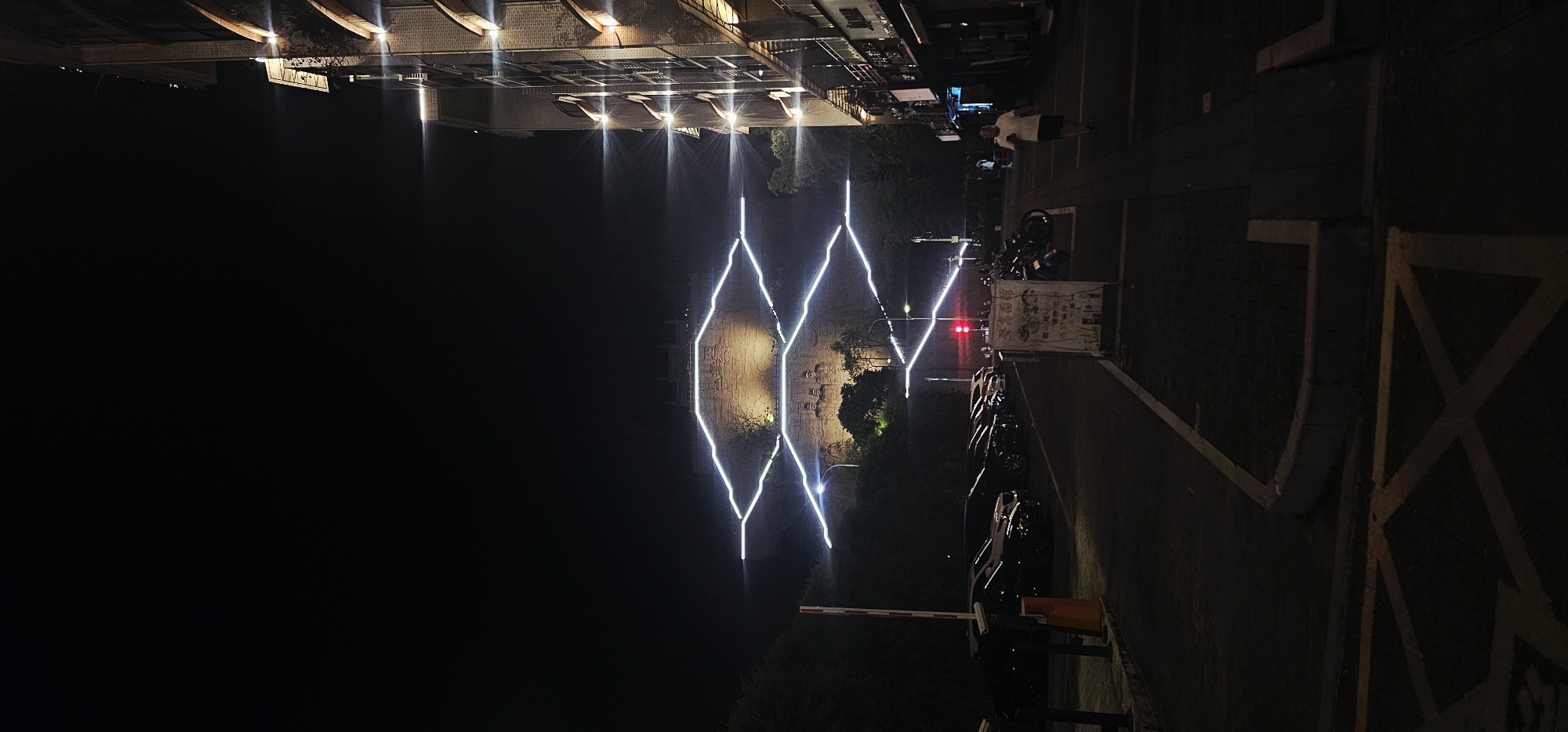 The image depicts an outdoor scene at night, likely in Beibei, China, featuring a building facade with illuminated windows and architectural lighting on the right side. In the background, a hillside is visible with a large carved stone monument or structure illuminated by bright white lights that form a zigzag pattern across its surface. Below the illuminated hillside, there appears to be a parking lot with several dark-colored cars parked in a row. A red traffic light is visible in the middle ground. On the right side of the image, a sidewalk runs alongside the building, and a lone person is walking on it, wearing a white shirt and dark shorts. The overall atmosphere is dimly lit, with the primary light sources being the building's illumination, the lights on the hillside monument, and the headlights of vehicles, creating a nocturnal urban or semi-urban environment.
