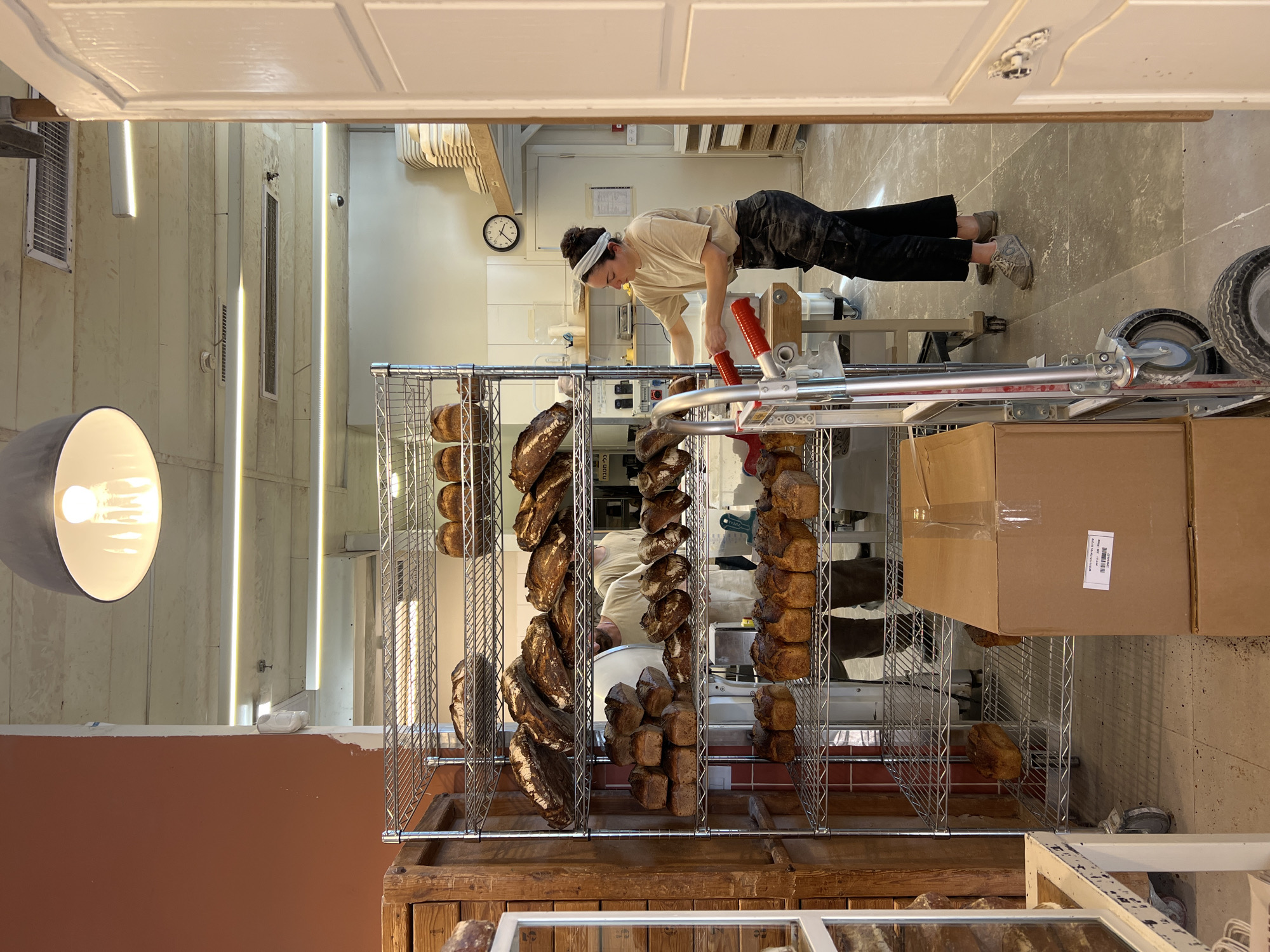 The image captures the interior of a commercial bakery in Tel-Aviv, Israel. The scene is dominated by two large, multi-tiered metal wire racks positioned on the left, extensively filled with various types of freshly baked bread loaves, including large sourdough rounds and rectangular sandwich loaves.
On the right side, a female baker is actively working. She wears a beige short-sleeved t-shirt, dark cargo pants, and a light-colored headband holding her hair in a bun. She is positioned at a wooden work table, wearing red gloves, and her hands are engaged with a white container. In the mid-ground behind the bread racks, two male figures are partially visible, both wearing light-colored shirts, also appearing to be working within the bakery.
In the immediate foreground, lower right, two brown cardboard boxes are stacked, the top one bearing a white label with Hebrew text "לחם מחמצת" (Sourdough Bread) and a barcode. A silver hand truck with red handles and black wheels is partially visible to the left of the boxes. The floor is light grey, showing signs of flour or dust.
The ceiling features exposed concrete, linear LED light fixtures, air vents, and a large, dark grey industrial pendant light fixture illuminating the foreground. A black-and-white analog wall clock on the back wall indicates a time of approximately 1:50. Stacks of white plates or bowls are visible on shelves in the far-right background, along with a small, white dome security camera mounted on the back ceiling. A partially open white, paneled door frames the right edge of the image.