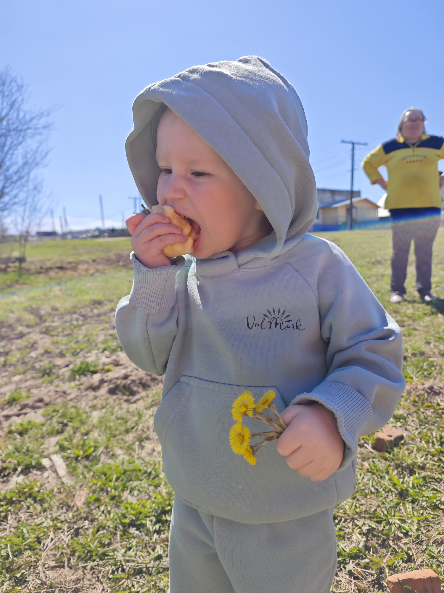 A young child stands outdoors in a field in Фоки, Russia, wearing a light blue-gray hooded sweatshirt and matching pants. The sweatshirt has the word "Vol Mark" printed on the chest. The child is eating something from their hand and holding a small bouquet of yellow flowers in the pouch of the sweatshirt. Behind the child, an adult stands with their hands on their hips wearing a yellow shirt with navy accents. The sky is blue, and the ground is a mix of grass and dirt. A building and utility poles are visible in the background.