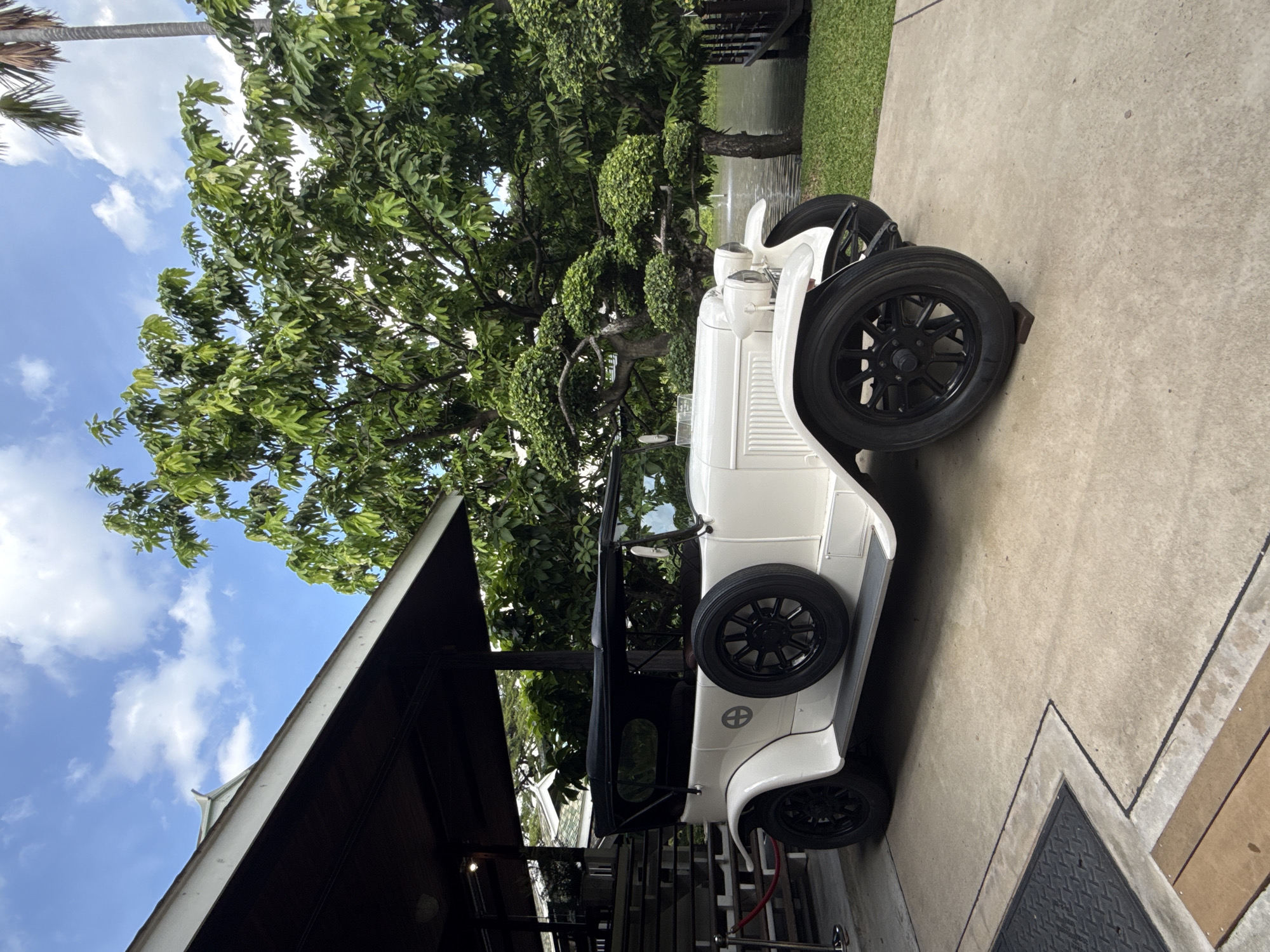 A white vintage car is parked on a concrete surface. The car has a black convertible top, black spoked wheels, and a spare tire mounted on the left rear fender. It is parked under a covered patio area with a dark brown roof supported by dark posts. To the left of the car, a red velvet rope barrier is visible, with steps leading up to the patio. Beyond the car, a lush green garden with trees, including some topiaries, and a body of water with a fountain can be seen. The sky is blue with scattered white clouds. The scene appears to be outdoors, possibly at a restaurant or hotel in Bangkok, Thailand, given the tropical vegetation. There are no people visible in the image.