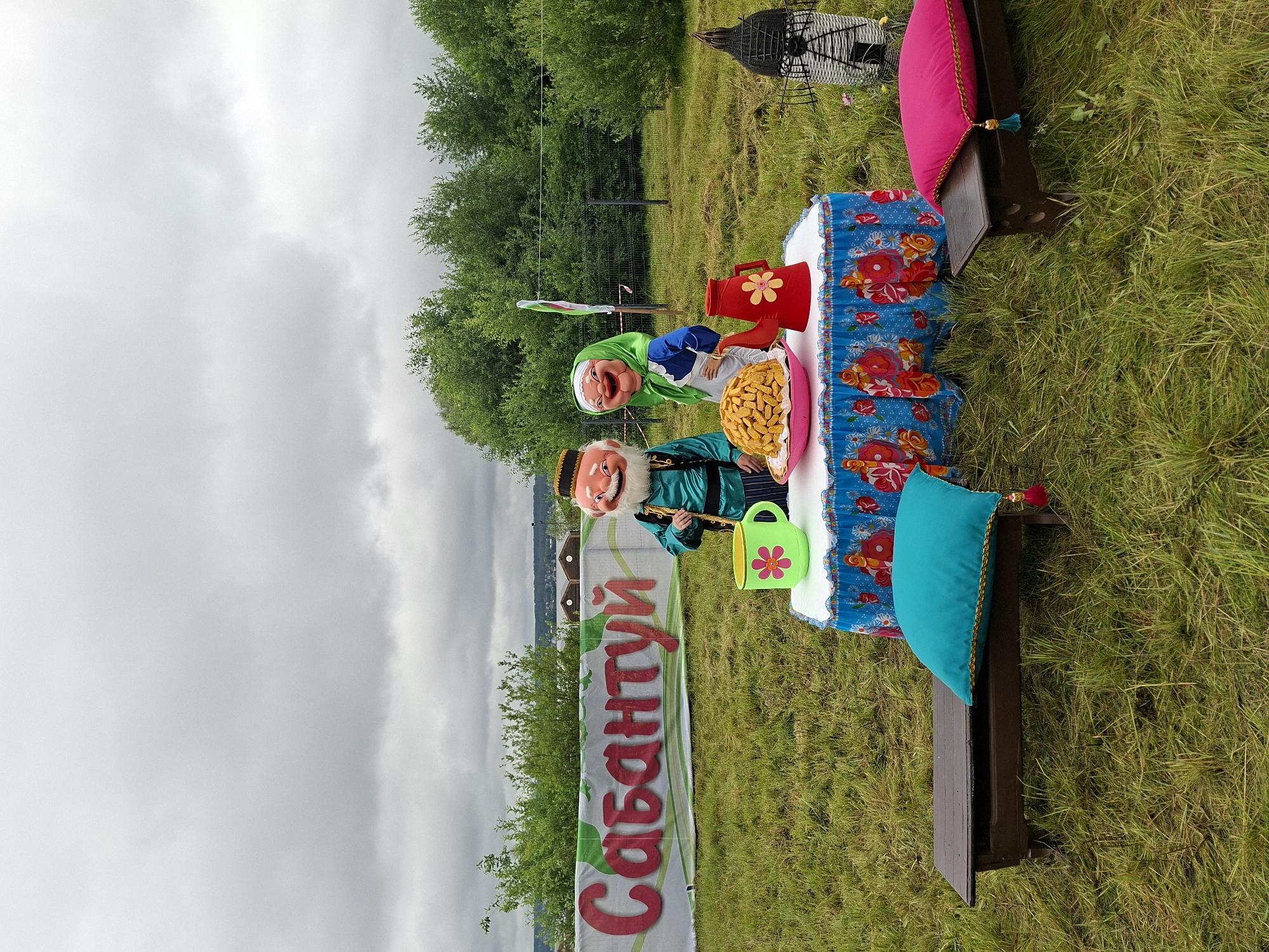 The image depicts an outdoor scene with a banner that reads "Сабантуй" (Sabantuy) in large red letters. Two costumed figures, a man and a woman, are standing near a table set up on the grass. The man is wearing a green robe and a patterned cap, and the woman is in a blue dress and a green headscarf. On the table, there is a large pink bowl filled with what appears to be baked goods, a red teapot with a floral design, and a green mug with a pink flower. The table is covered with a blue cloth featuring a floral pattern. Two wooden benches, one with a turquoise cushion and the other with a pink cushion, are placed on either side of the table. In the background, there are trees and a cloudy sky. The Sabantuy is a traditional Tatar and Bashkir festival, celebrated in various regions of Russia, including Votkinsk, Udmurtia, which is located on the Kama River. The scene appears to be set up for a festive event.