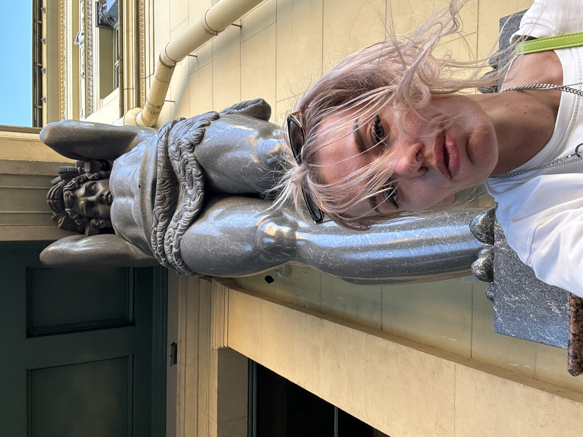 This is a vertical, eye-level, close-up selfie taken outdoors in Saint Petersburg, Russia. The main subject is a young woman with blonde hair, wearing sunglasses pushed up on her head and a white t-shirt with a green strap and silver chain necklace. She is looking directly at the camera with a neutral expression. To her left is a large, grey stone sculpture of a reclining male figure. The sculpture appears to be made of granite and depicts the muscular body of a man. The woman is positioned such that her face is in the foreground, and the sculpture occupies the midground and background. Behind the sculpture, a light-colored building with ornate architectural details and a cream-colored pipe are visible. The sky is a clear, bright blue, suggesting a sunny day. The overall scene has a classical or historical ambiance due to the presence of the sculpture and the architecture. The image captures a moment of tourism or exploration, with the woman posing playfully with the artwork. The time of day appears to be daytime due to the bright lighting and clear sky.