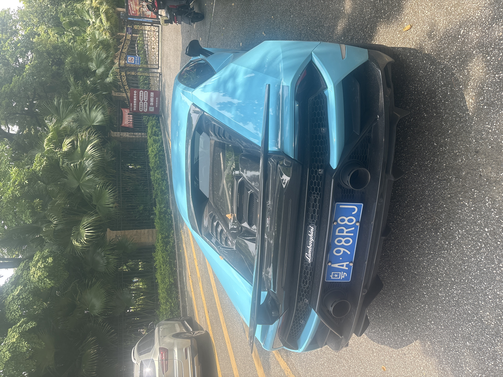 A bright blue Lamborghini sports car is parked on an asphalt road. The car's rear is facing the camera. The license plate is visible and reads "A·98R8J". The car has a large rear spoiler. Behind the car, there is a black metal fence, a gate with ornate gold detailing, and lush green foliage including palm trees. To the left of the Lamborghini, a silver SUV is parked. Further in the background, signs with Chinese text are visible near the gate. A red and yellow sign on a red stand is also present near the gate. A motorcycle is parked to the right of the gate. The environment appears to be a residential or gated community entrance within Nanhai District, China.