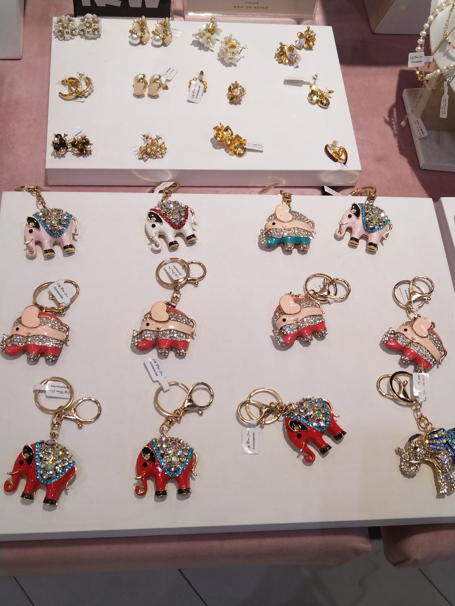 The image displays a close-up, eye-level view of an assortment of costume jewelry displayed on white risers. The jewelry is primarily gold-toned and features rhinestones, enamel, and pearl accents. In the foreground, several elephant-shaped keychains are arranged in rows. These keychains are adorned with colorful enamel and rhinestones. Above the keychains, on a higher riser, are various pairs of earrings, including floral designs, hoops, and stud earrings. The background is a soft pink fabric, likely a display cloth. The lighting is bright and even, suggesting an indoor retail environment. There are no people visible in the image. The jewelry appears to be new and well-maintained. The scene is consistent with a display in a jewelry store or market stall.