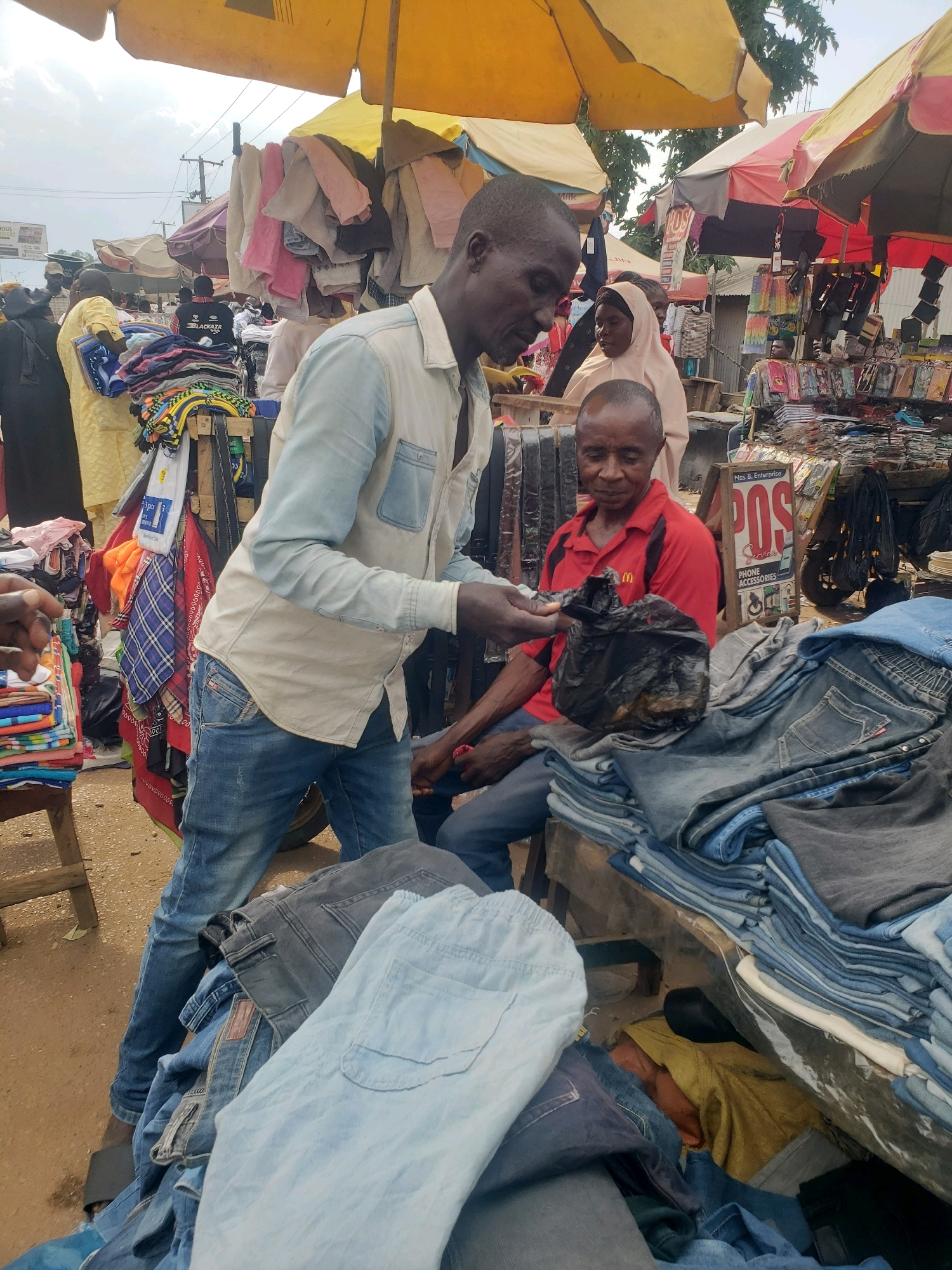 In Sabon Tasha, Nigeria, a street market scene unfolds under a yellow canopy. A man wearing a light blue collared shirt and blue jeans stands at a stall filled with folded denim. He is handing a black plastic bag to another man seated across from him, who is wearing a red polo shirt. The seated man appears to be inspecting the contents of the bag. Piles of folded jeans, in various shades of blue and grey, are displayed on a wooden table in front of the seated man. In the background, other market stalls are visible, with vendors and shoppers milling about. Colorful textiles, clothing racks, and various merchandise are spread out, suggesting a bustling commercial area. The ground is dusty, indicative of an outdoor market setting. The sky above is overcast.