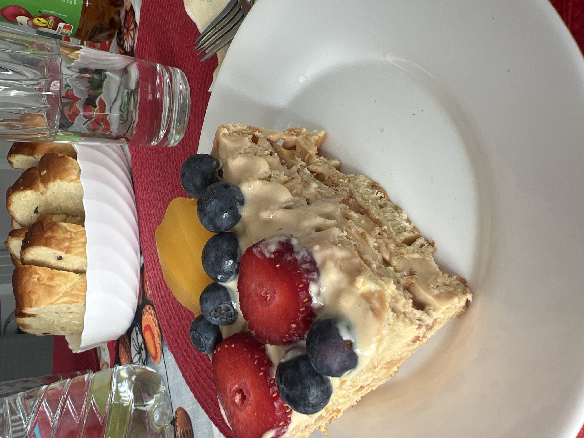 A slice of layered cake topped with cream, strawberries, blueberries, and a yellow fruit slice sits on a white plate. A stack of sliced bread is adjacent to the cake, contained in a white paper wrapping. A glass of liquid is also present. A fork is positioned near the cake on a red patterned placemat. The setting might be a private residence or a café, but no information is available to determine that this image was taken in Yambol, Bulgaria.