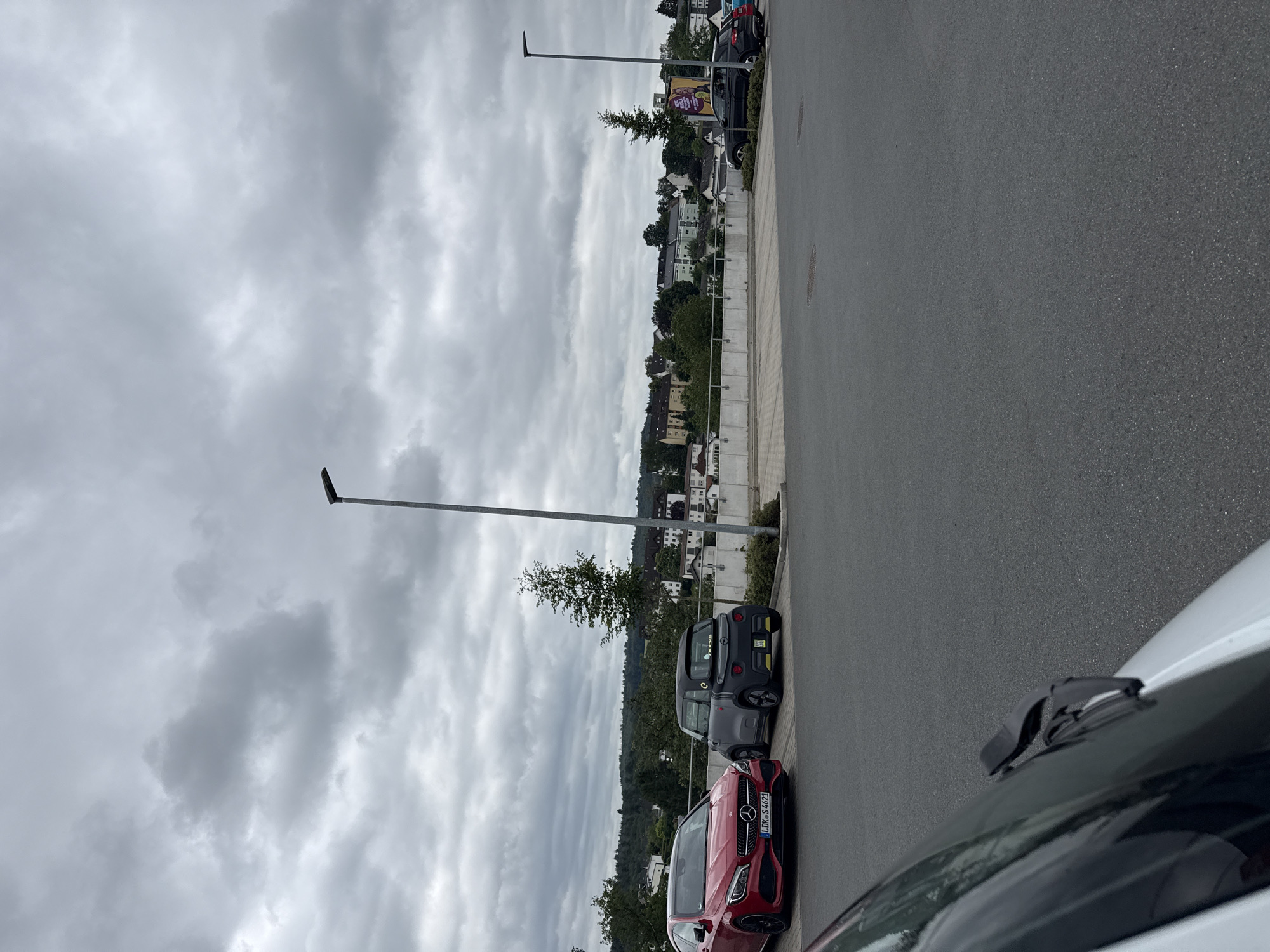 The image shows a parking lot in Braunfels, Germany, under a cloudy sky. In the foreground, a portion of a white vehicle's windshield and wiper blades is visible, angled towards the right. To the left of the image, a red Mercedes-Benz CLA-Class vehicle is parked, displaying the license plate "LDK S 4621". Next to it, a small, dark gray electric car with yellow accents is parked. In the background, a line of trees and buildings forms a cityscape on a hillside. A lamppost stands to the right of the cars, and another lamppost is visible further in the background near a billboard. The terrain is paved with asphalt.