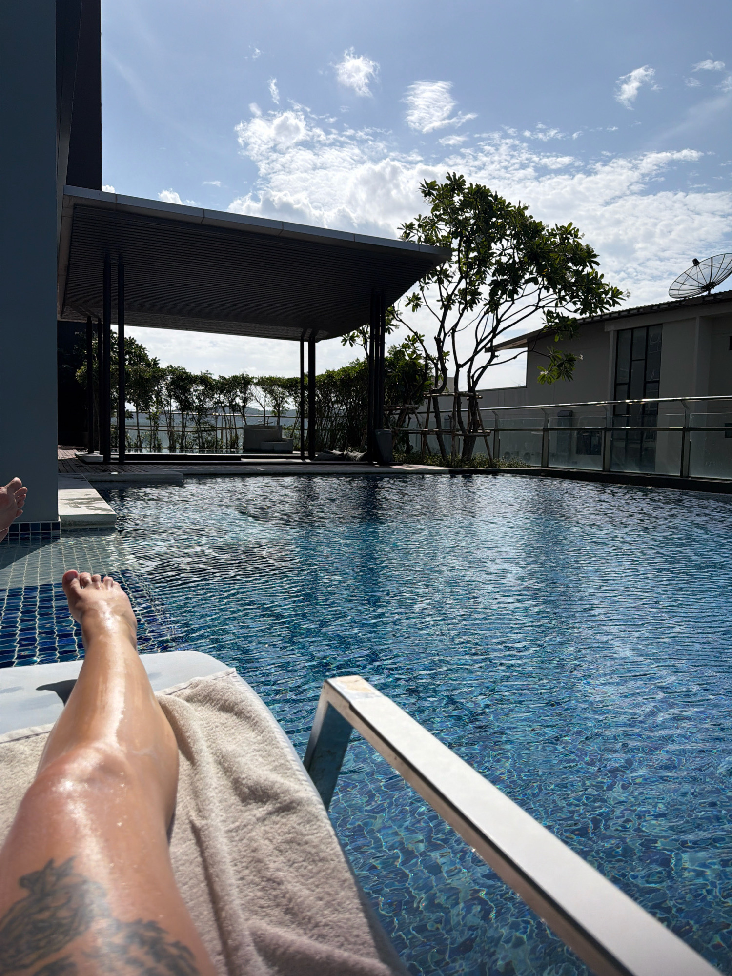 A first-person perspective shot shows a person's legs and feet resting on a towel on a lounge chair next to a swimming pool. The pool water is a deep blue and has ripples from the sunlight. In the background, there is a covered seating area with supporting pillars, lush green trees, and a building with a satellite dish on the roof. The sky is a bright blue with scattered white clouds, indicating a sunny day. The scene appears to be a resort or hotel in Hua Hin City Municipality, Thailand, likely during daytime hours.
