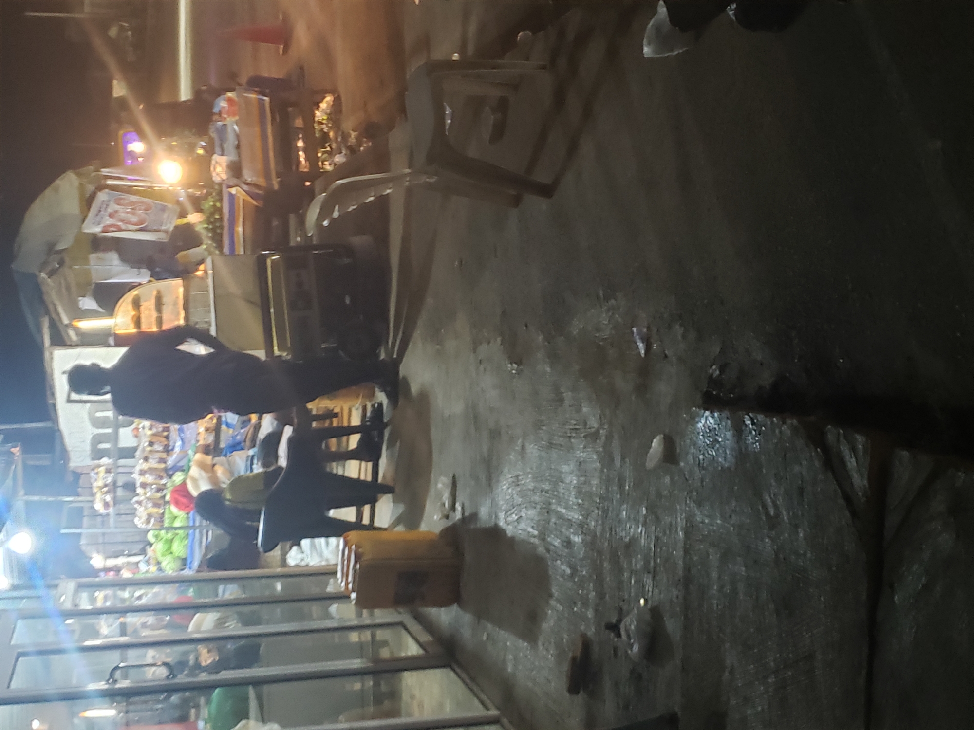 The image depicts an outdoor night market or street scene in Ibadan, Nigeria. The time of day is clearly night, with various artificial light sources illuminating the area, casting reflections on the wet ground. The ground appears to be concrete or asphalt, showing signs of wetness, possibly from rain or spilled liquids.
Several people are present, indicating activity. One individual, wearing dark clothing, stands with their back to the viewer, facing a market stall. Another person is seated on a black plastic chair within the stall area. Other figures are partially visible further in the background and within the stalls.
The scene is bustling with market elements. There are multiple stalls or stands featuring various goods, including what appears to be fresh produce like green and red items (possibly vegetables or fruits) displayed in a refrigerated or glass-fronted case. Above these, strings of small, packaged items are visible, possibly snacks or other goods. A white plastic chair is also visible on the wet ground. In the foreground, a yellow jerrycan or container is placed on the ground, with the word "CASH" partially visible on its side. A white package with red text, possibly "SAUSAGES," lies on a counter in one of the stalls. A red traffic cone is seen near the background. The bright lights from the stalls create strong illumination in some areas and deep shadows in others, contributing to the typical atmosphere of a lively night market.