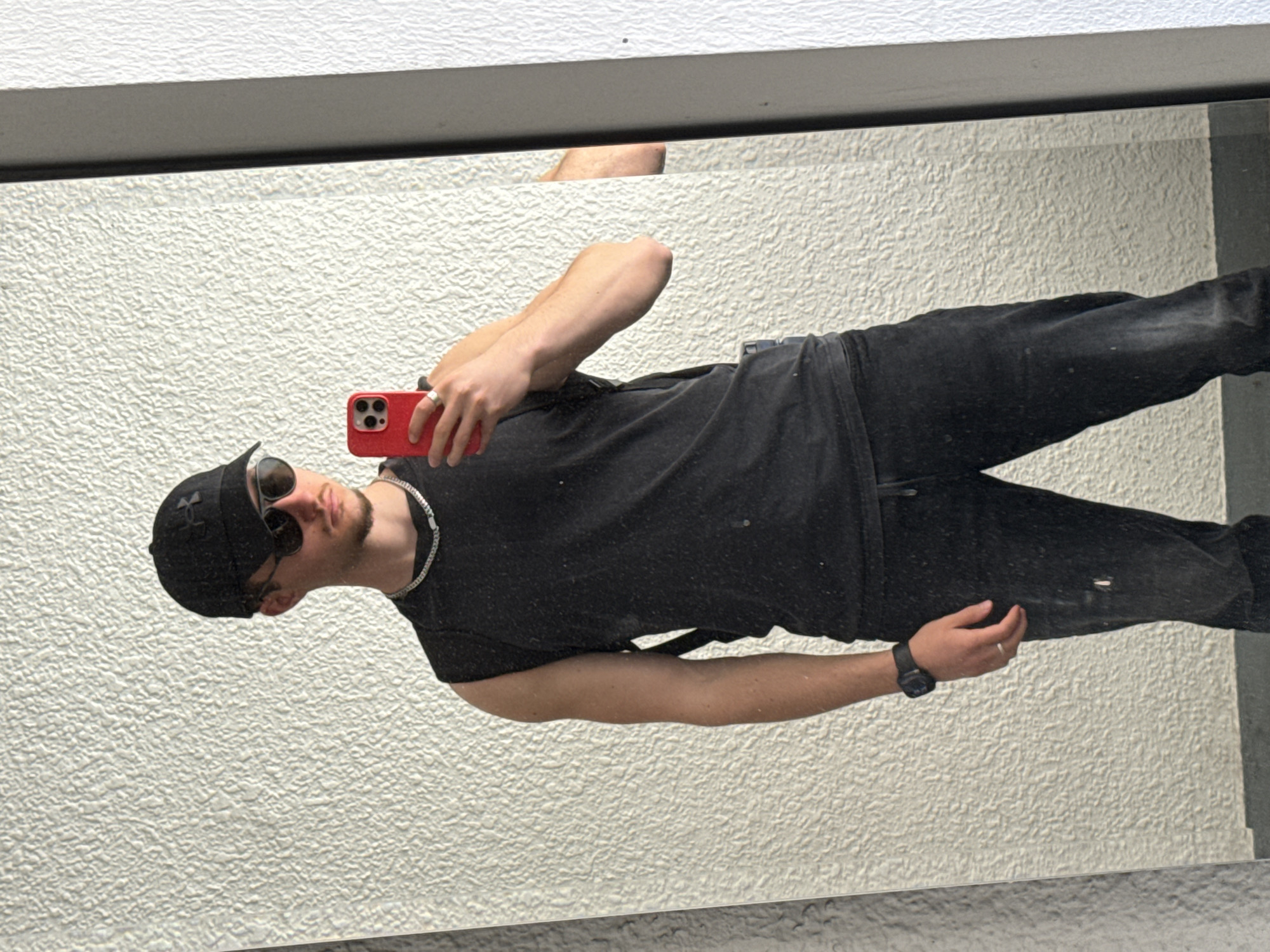 A male is reflected in a mirror, holding a red smartphone. He is wearing a black baseball cap, sunglasses, a black t-shirt, black jeans, and a watch. He also has a silver necklace and a ring. The mirror reflects a white textured wall. Above the mirror, part of a hand is visible. The location is not identifiable from the image.