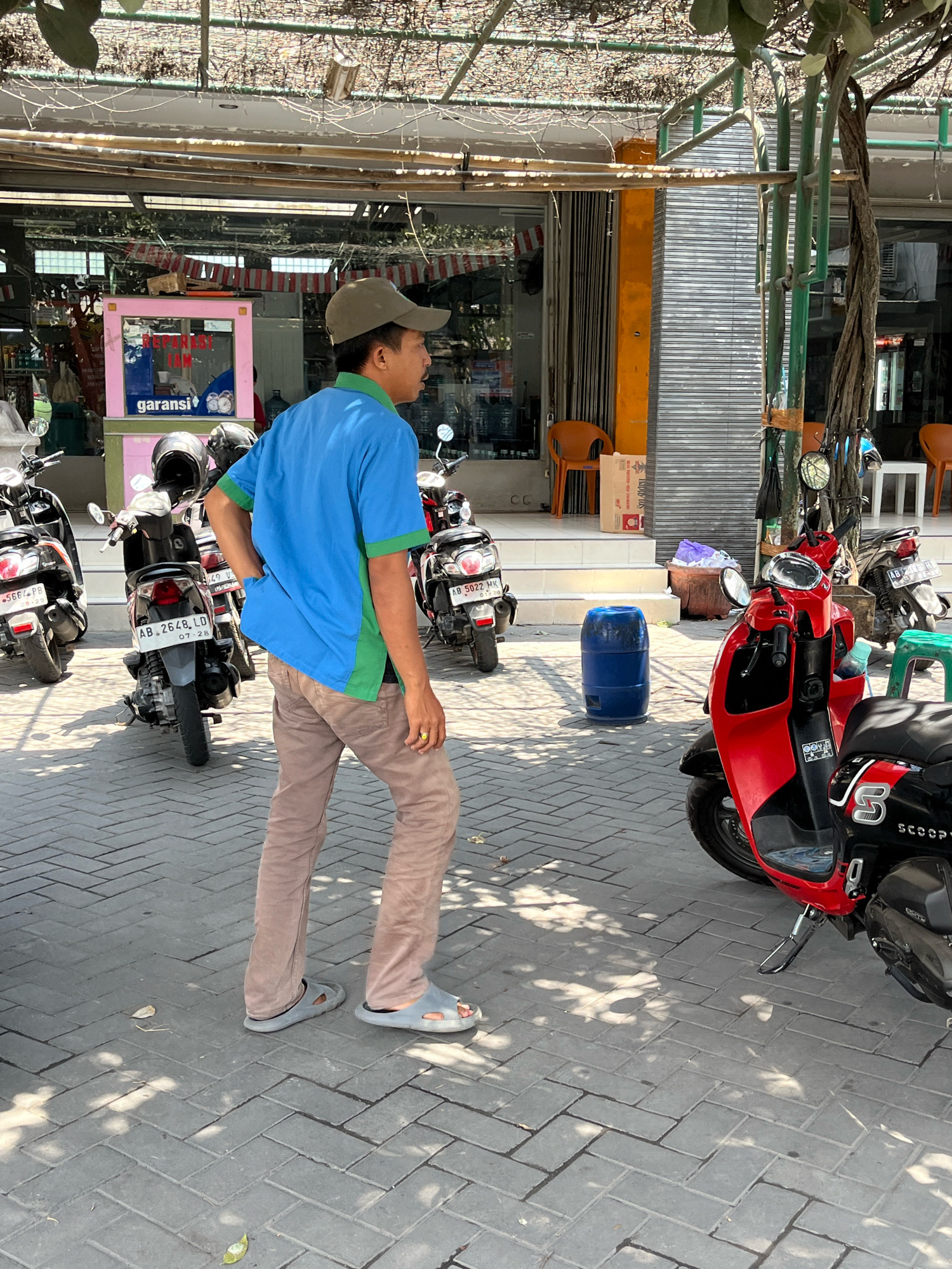 The image depicts a street scene in Banguntapan, Indonesia, likely a commercial area. A man wearing a blue shirt with green accents, brown pants, and a green cap is standing in the foreground, facing away from the camera and towards a row of shops. He is wearing gray slip-on sandals and has a gold ring on his right hand. Behind him, several motorbikes are parked on a paved sidewalk. The shops have metal roll-up doors, and some appear to be open, with displays visible inside. There are orange chairs placed outside one of the establishments. A large blue plastic barrel stands on the sidewalk near the motorbikes. In the mid-ground, a bright red scooter is parked prominently, with a black and silver scooter behind it. The environment is bright and sunny, with shadows cast by overhead structures and trees.