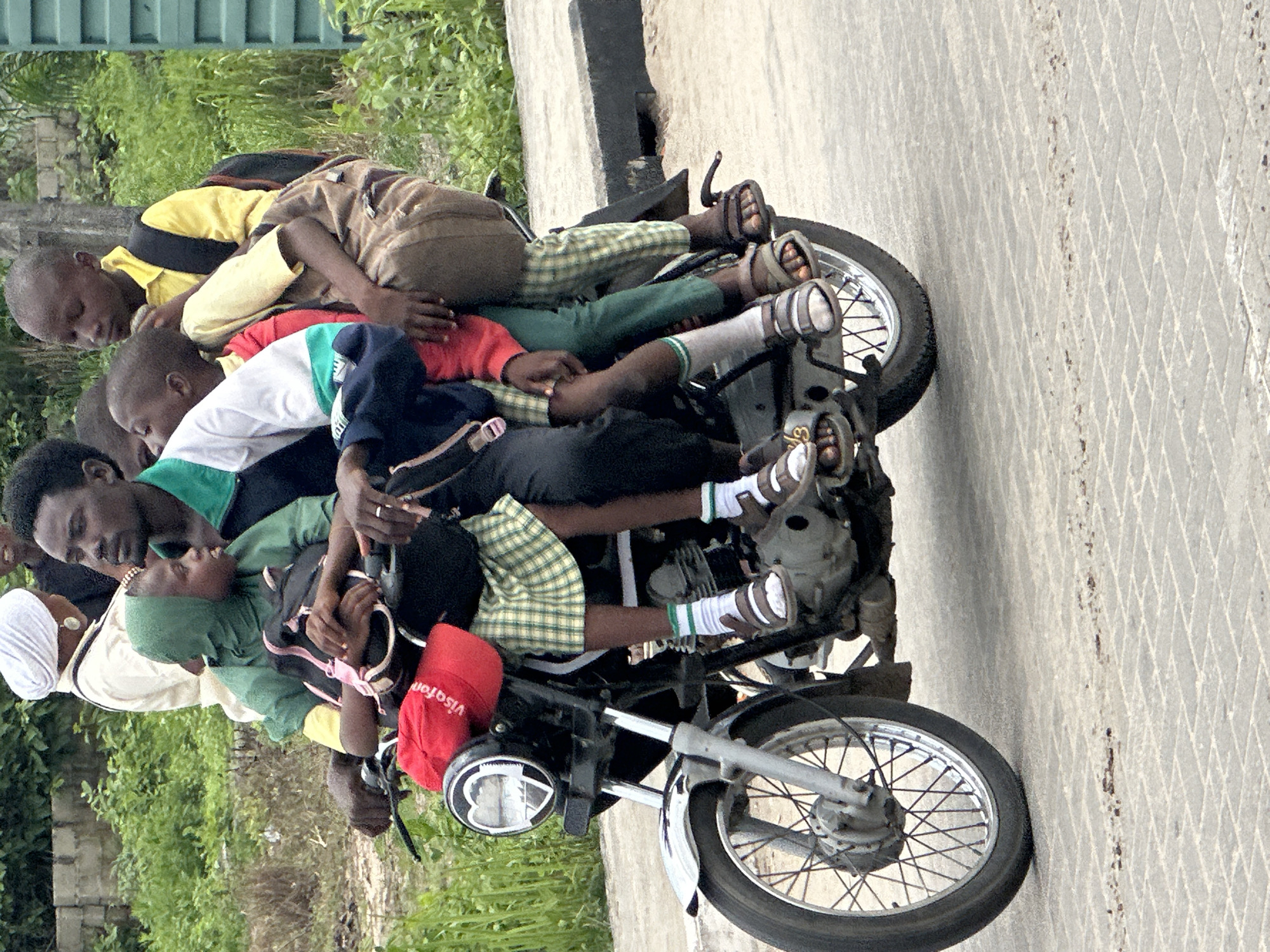 This media file depicts an extremely overloaded motorcycle carrying at least nine people, including adults and numerous children, in an outdoor setting in Badagry, Nigeria.
The subjects are tightly packed onto the single motorcycle. An adult male with dark skin and a green shirt appears to be at the front, possibly driving, while holding a young child in a green headscarf. Behind him, another adult, partially obscured, wears a white headscarf and light-colored clothing. The rest of the motorcycle is covered by children. One child in a yellow and black striped shirt and brown backpack is lying across the rear, with another child in a red shirt and green pants immediately beside them. Further down, a child in a white, green, and blue striped shirt is visible, and another child is seen wearing green plaid shorts, white socks with green stripes, and sandals. Several other children are nestled amongst the group, making a precise count challenging.
The scene takes place on a paved surface, likely a road or path, which features a distinctive diamond pattern. Lush green vegetation, including bushes and grass, lines the side of the road, with a concrete wall or barrier visible behind it in some sections. The bright, clear lighting suggests it is daytime.
The most notable detail is the severe overloading of the motorcycle, far exceeding its intended capacity and posing a significant safety concern. No occupant is seen wearing a helmet or any other protective gear. Despite the precarious situation, the individuals generally appear calm. The only visible text is the word "Visafon" printed on a red baseball cap worn by one of the children.