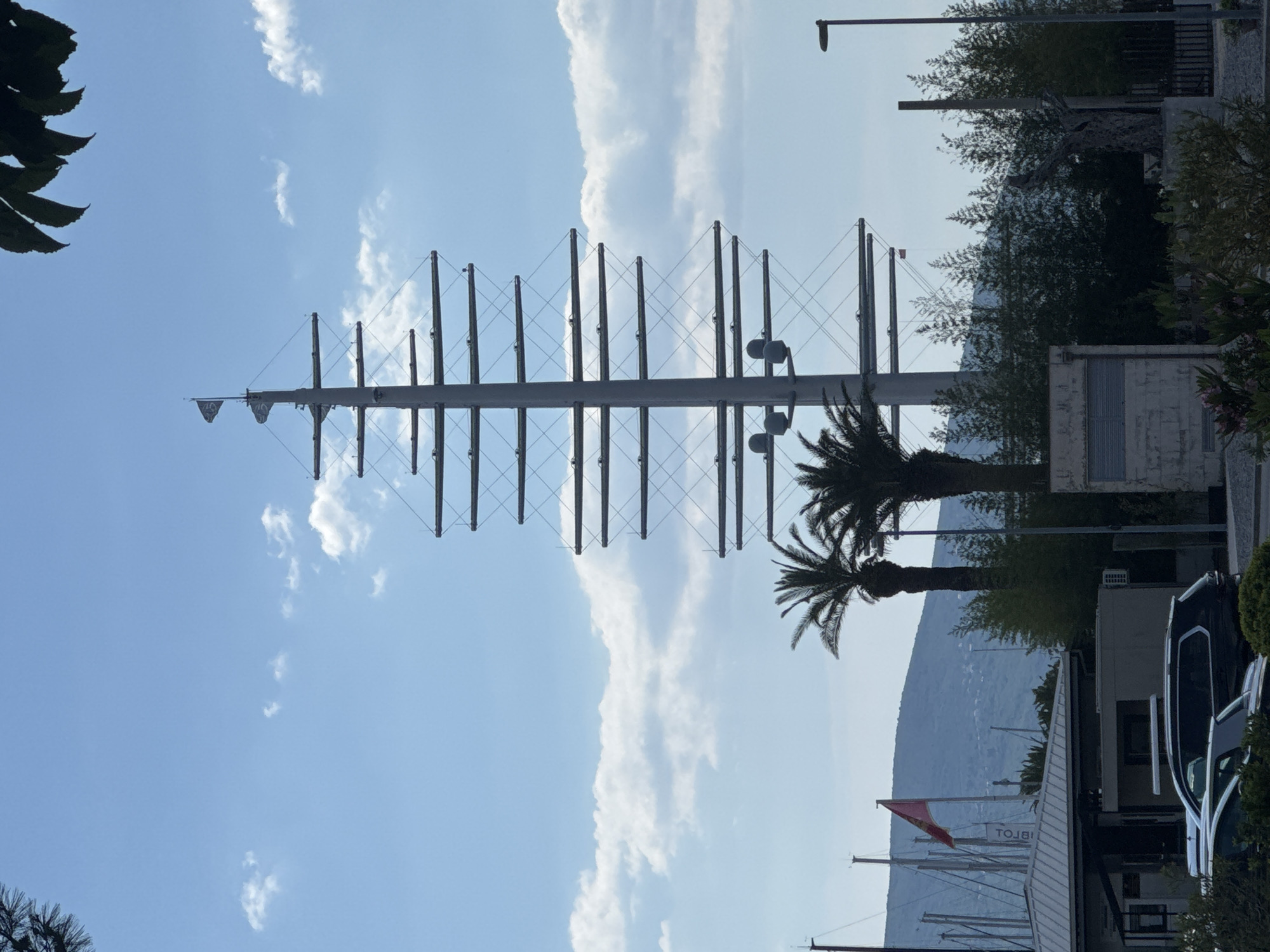 The image shows a tall, metal sculpture resembling the mast of a sailing ship, situated in Tivat, Montenegro. The sculpture is comprised of a central vertical pole with horizontal crossbars supporting numerous evenly spaced vertical posts connected by cables. Two palm trees partially obscure the base of the structure. Behind the trees, a mountain is visible in the distance. To the right of the sculpture and trees are various buildings, a street lamp, and a parked van. The sky is blue with scattered white clouds.