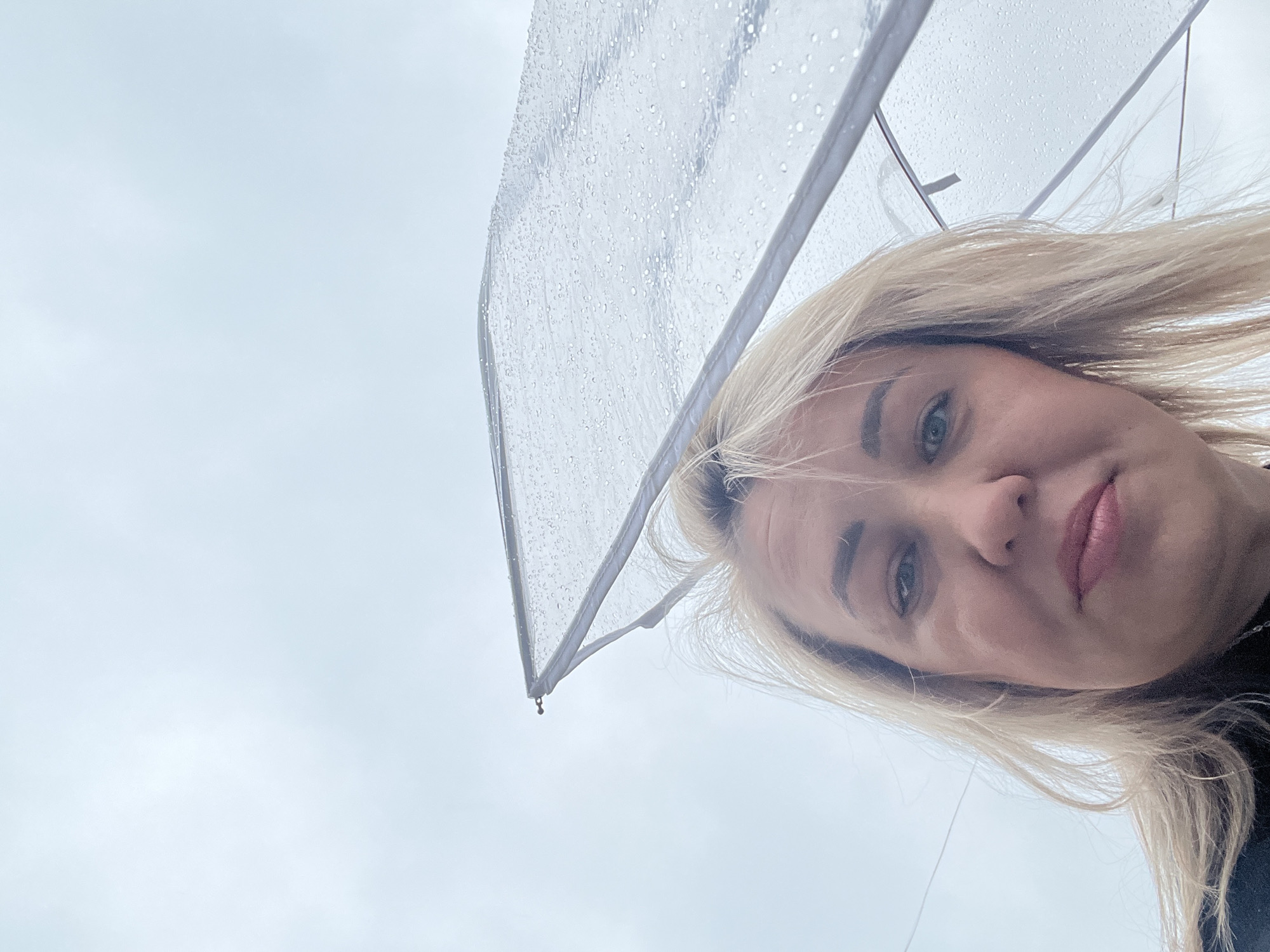 A woman with blonde hair, likely in her 30s or 40s, is holding a clear umbrella. The umbrella has water droplets on it. The sky is overcast. No specific details confirm the location as Chaikovsky, Russia.