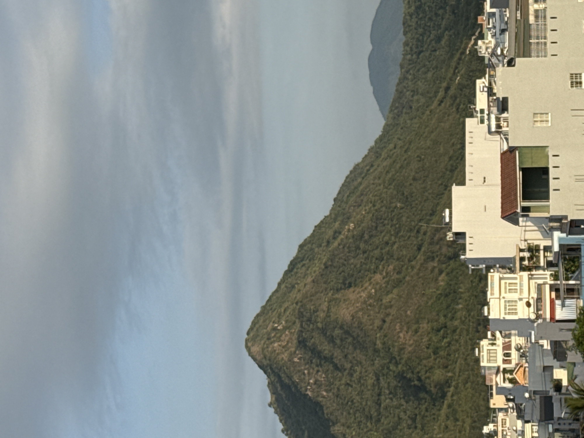 The image shows a view of Bac Nha Trang Ward, Vietnam, with a prominent green, tree-covered mountain dominating the background. In the foreground, a cluster of multi-story buildings with white and light gray facades, some featuring balconies and windows, are visible. The sky above is a mix of light blue and overcast gray clouds, with some wispy streaks of clouds. The lighting suggests it might be late afternoon or early morning. There are no people or specific actions depicted in the image. The scene is a typical urban landscape set against a natural mountainous backdrop.