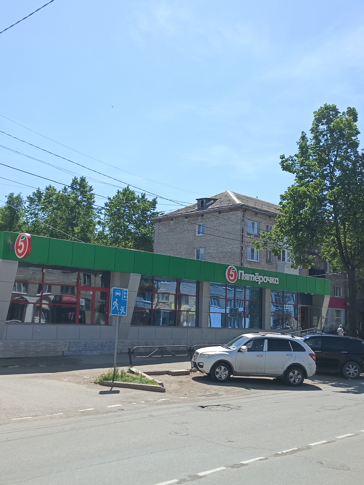 The image shows a "Pyaterochka" supermarket in the city of Chaikovsky, Russia, recognizable by its green and red signage. The building is a multi-story brick structure, likely a residential apartment building integrated with the supermarket storefront. A white SUV is parked in front of the store, and a black car is parked behind it. A sign indicating a pedestrian crossing with a bus stop is visible. The sky is clear and blue, and there are trees in the background. Overhead power lines are also visible.
