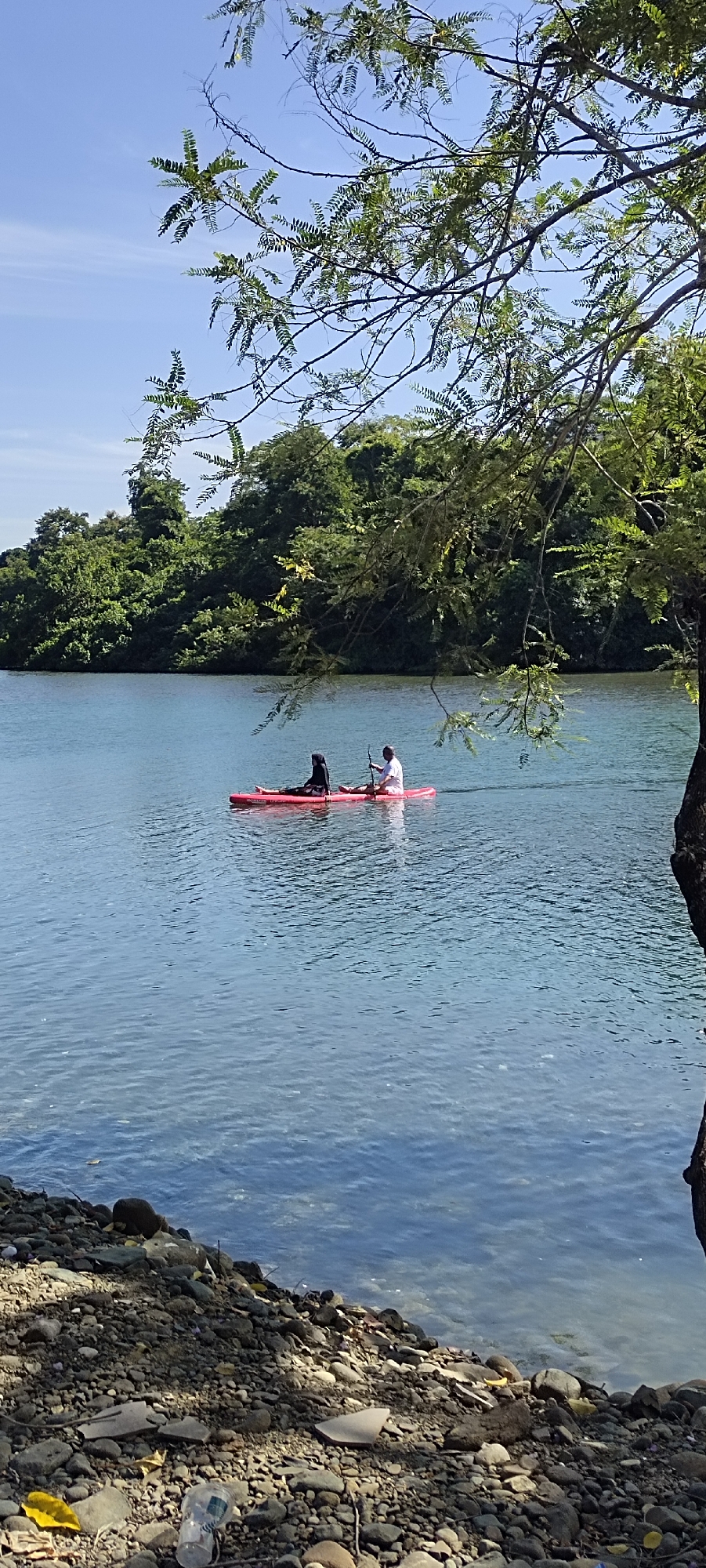 Two individuals are paddleboarding on a tranquil, clear blue river. One person, dressed in a black hijab and attire, is seated towards the front of the red paddleboard, while the other, in a white shirt and shorts, is seated towards the back, holding a paddle. The river reflects the bright blue sky and is bordered by lush green trees on the opposite bank. The foreground features a rocky shoreline with scattered debris, including a yellow leaf and a clear plastic cup. A tree with green leaves partially obscures the view in the upper right corner of the image. The scene suggests a serene natural setting, possibly in the city of Lamseunia, Indonesia.