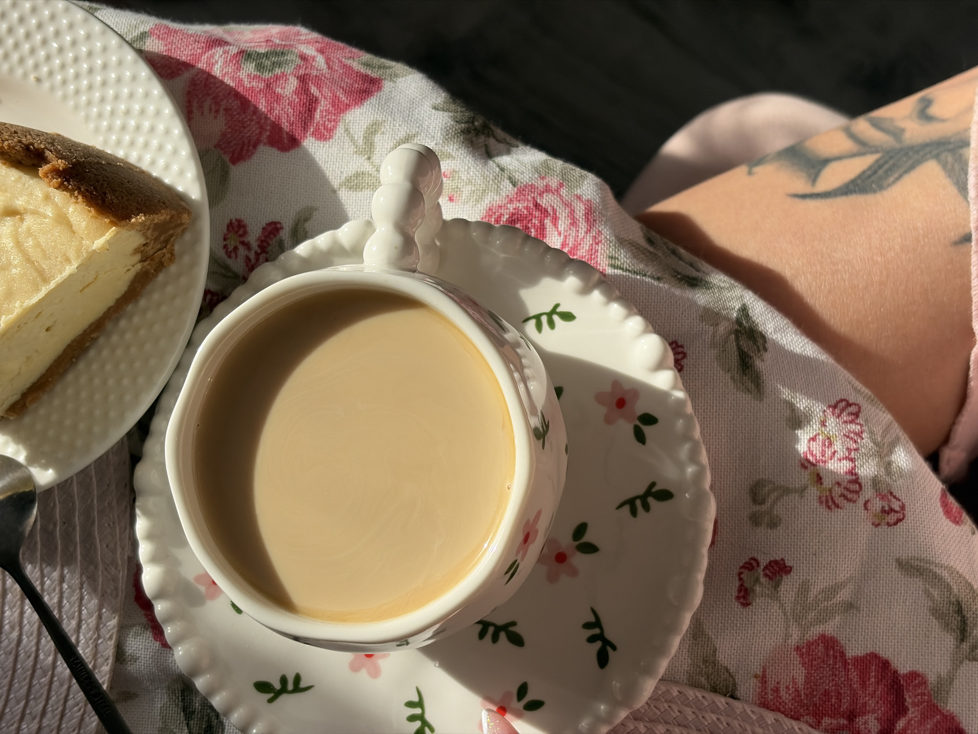 A high-angle shot depicts a cup of coffee on a saucer, a slice of cheesecake, and a tattooed arm. The cup, saucer, and plate are white with floral patterns. The coffee is light brown in color. The cheesecake has a golden crust and a creamy filling. A spoon is visible next to the plate. The floral tablecloth beneath the items adds a soft, decorative touch. The tattoo on the arm appears dark. There's nothing to indicate the location of Saint Petersburg, Russia.