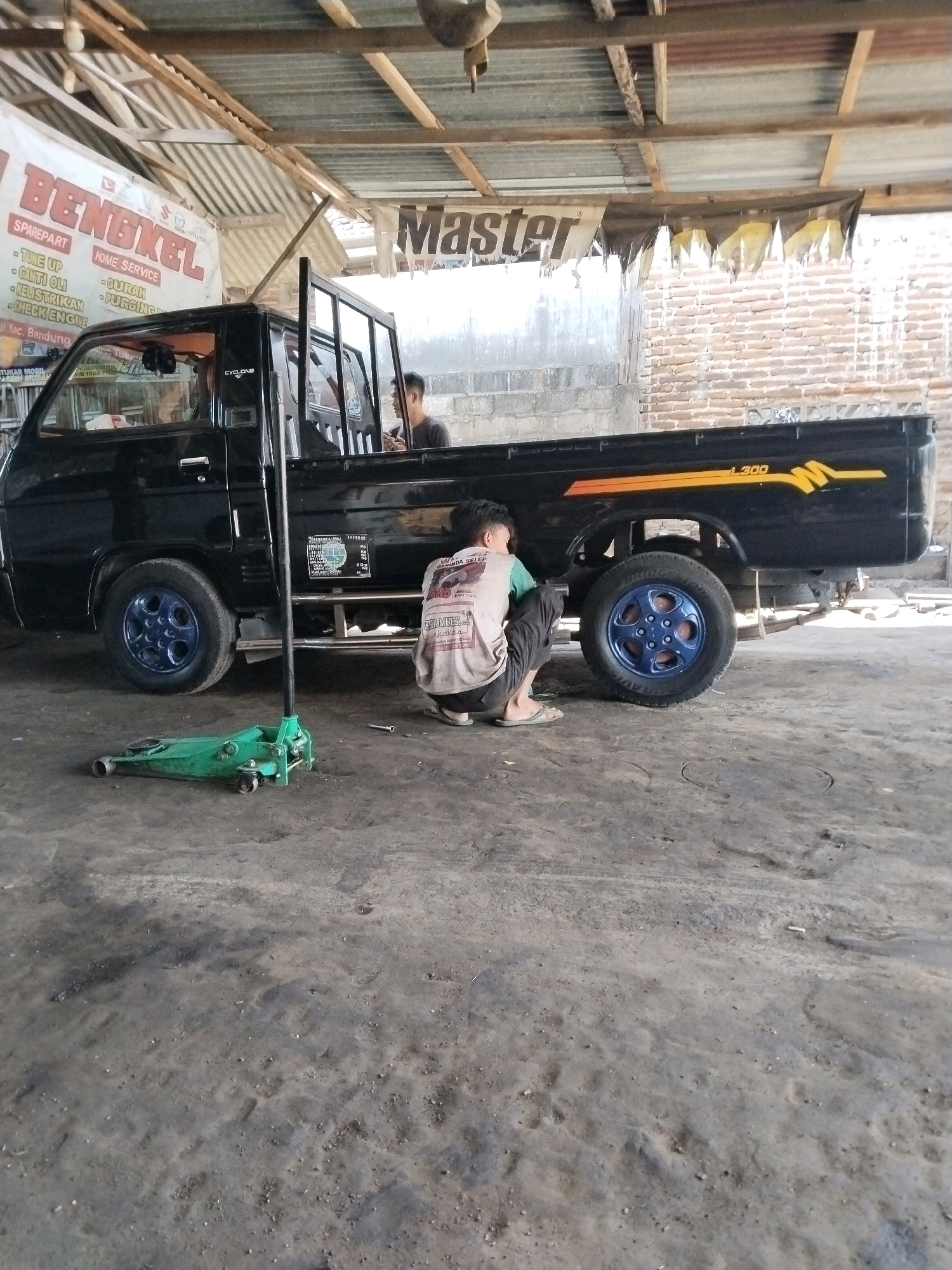 The image depicts a scene within a garage in Bandung, Indonesia. A black pick-up truck is parked on a concrete floor. A mechanic, wearing a light-colored shirt and dark pants, is crouched down beside the rear passenger-side wheel, appearing to be working on it. A green hydraulic jack is positioned on the ground near the wheel. In the background, a banner with the word "BENGKEL" (which means "workshop" in Indonesian) and "HOME SERVICE" is visible, along with another banner that reads "Master". The environment suggests an active workshop setting with exposed roofing and brick walls.