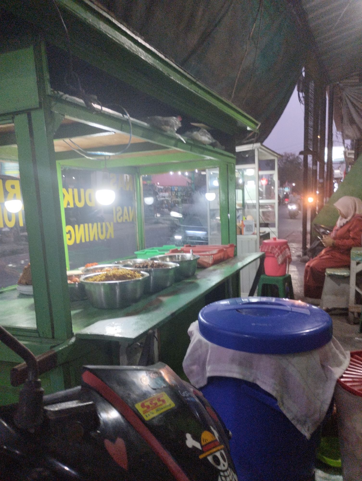 The image depicts a street food stall at dusk in Bekasi, Indonesia. The stall is illuminated by interior lights and streetlights, casting a warm glow on the food displayed in metal bowls. The stall is made of green painted wood and features a glass display case. Behind the stall, a woman wearing a hijab and a red garment sits on a stool, looking at her phone. In the foreground, a motorcycle is partially visible, adorned with stickers, including one with a skull and crossbones. The street appears busy with traffic, indicated by the headlights of passing vehicles. The overall scene conveys a typical evening atmosphere in a bustling urban area, where food stalls are a common sight.