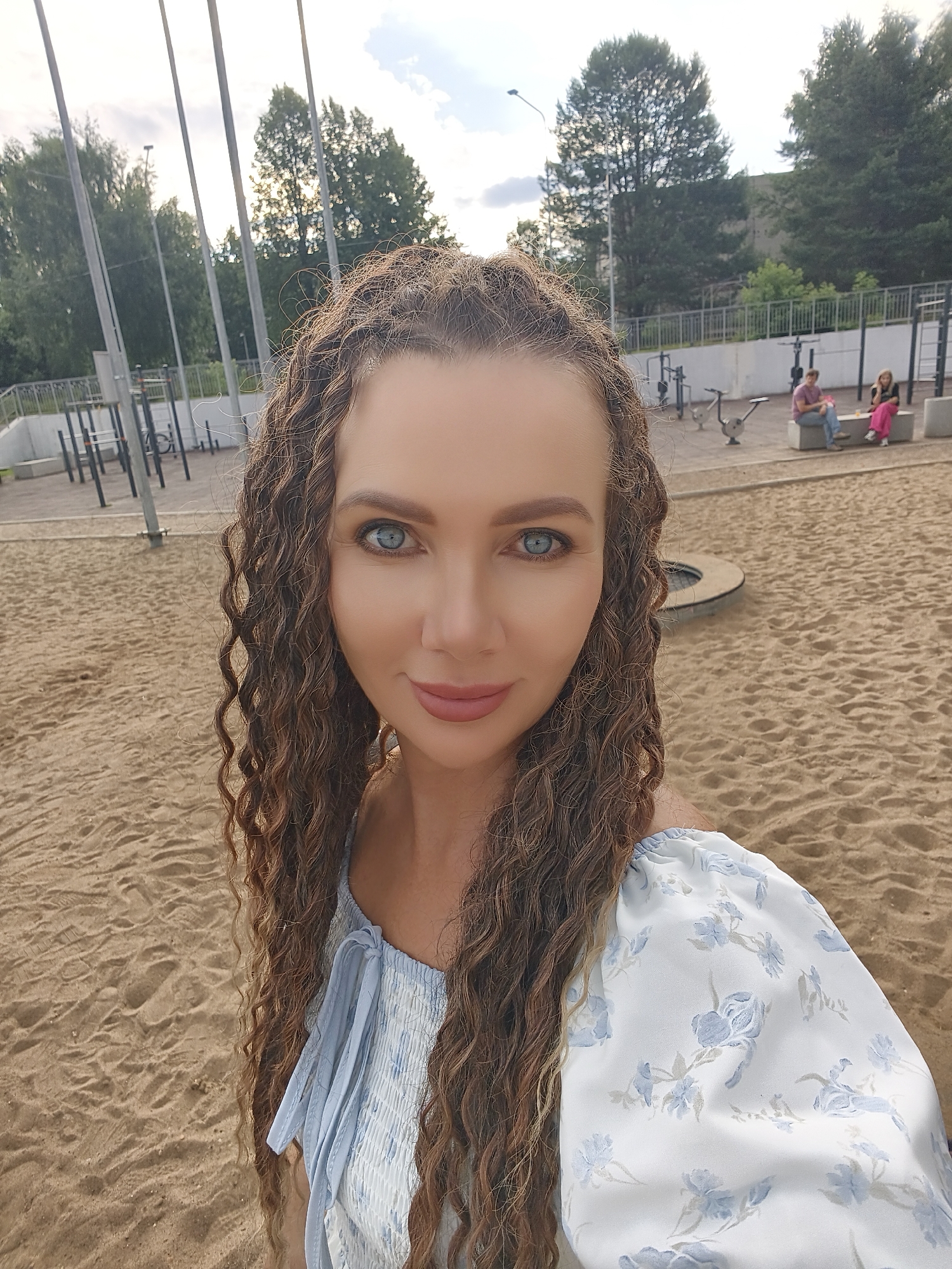 A woman with long, curly brown hair and blue eyes is taking a selfie in an outdoor sandy area, possibly a playground or park, with exercise equipment visible in the background. She is wearing a light blue off-the-shoulder top with a floral pattern. Two people are seated on a bench in the middle ground. The location appears to be Chaikovsky, Russia, based on its features.