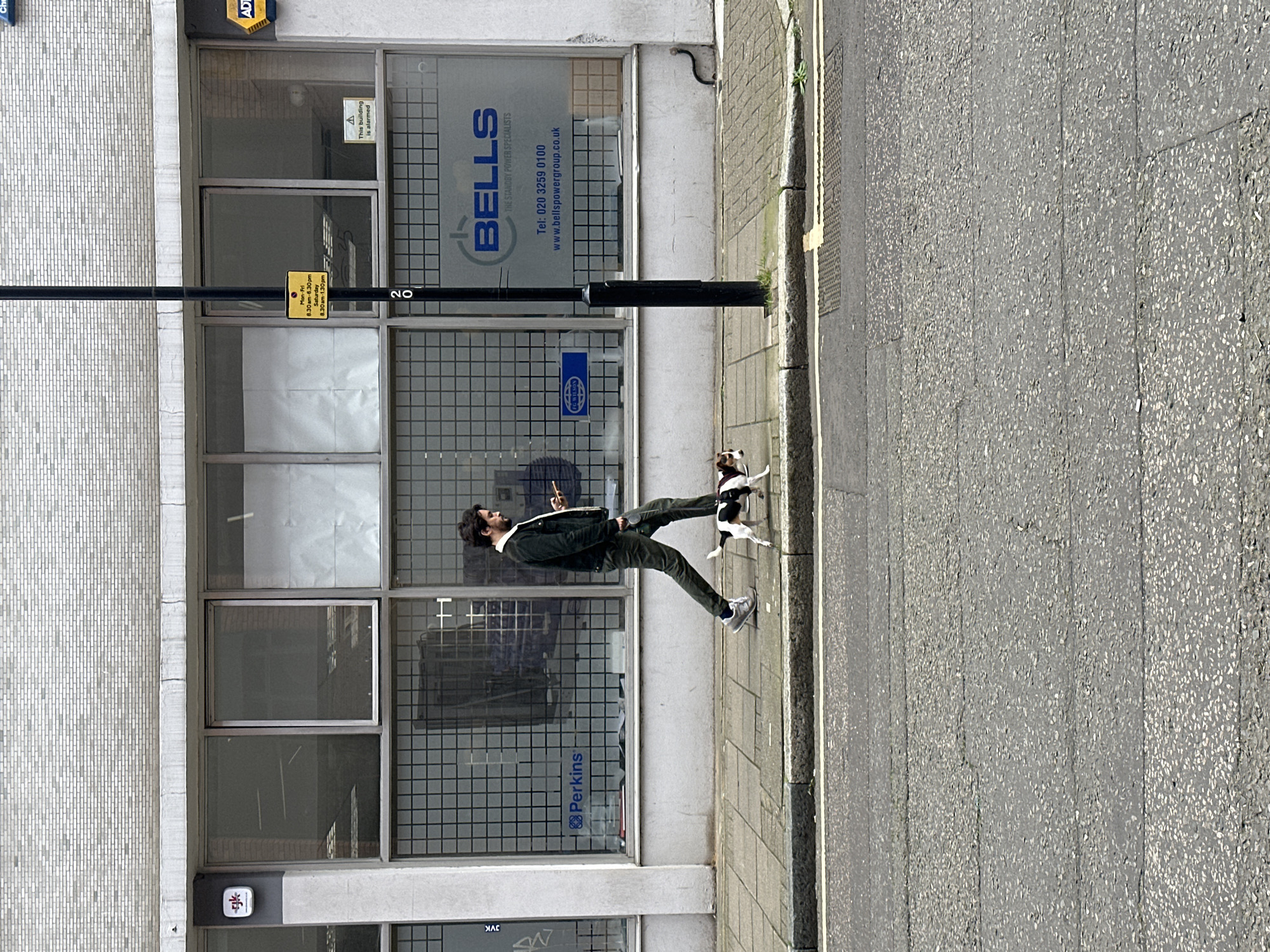 A man in a dark jacket and pants stands on a sidewalk in front of a building, looking at his smartphone. A small dog is near him. The building has large windows with signs for "BELLS THE STANDBY POWER SPECIALISTS" and "Perkins." Signs indicate the business hours are Monday-Friday 8:30am-6:30pm and Saturday 8:30am-1:30pm. The building number is 20. There is also a sign that reads "This building is alarmed". Another sign indicates FG WILSON. The location appears to be in the city of London, United Kingdom.