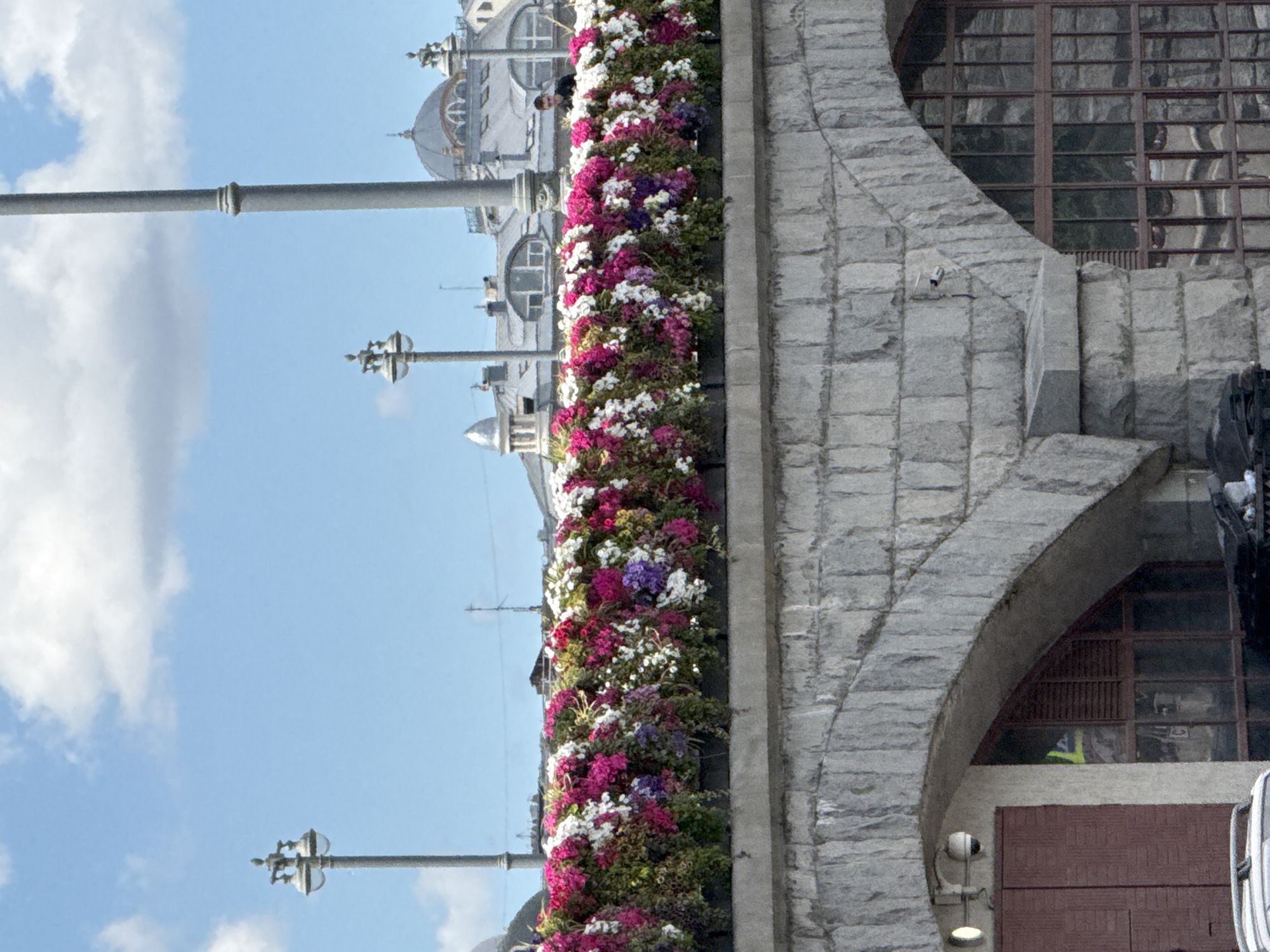 This media file shows a vertical composition featuring a building adorned with a lush vertical garden of flowers. The building's facade is constructed from large, rough-hewn stones, with a prominent arched entrance. Above the entrance, a large window with a grid-like pattern is visible, reflecting the sky and surrounding environment. The vertical garden overflows with a vibrant mix of pink, white, and purple flowers, cascading down the side of the building. In the background, parts of other architectural elements, possibly domes and spires, are visible against a bright blue sky with scattered white clouds. Several lampposts are also present, one in the foreground and others further back, suggesting an urban or public space. The overall scene appears to be outdoors, likely during the daytime given the bright lighting and clear sky. The atmosphere seems serene and perhaps festive, due to the colorful floral display. There is no discernible text in the image.