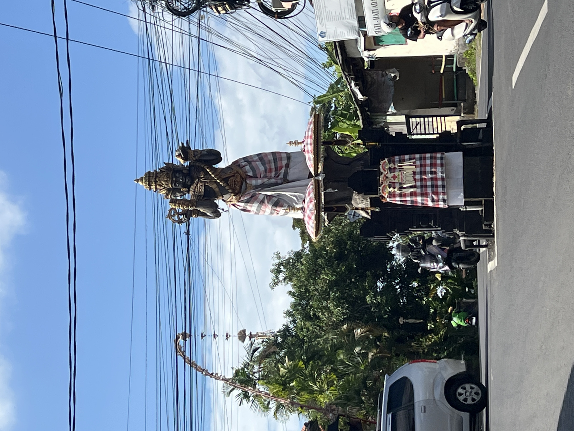A dark-colored statue with a golden headdress stands on a raised platform in the center of a street intersection in North Kuta, Indonesia. The statue has multiple faces and holds various objects in its hands. The platform is adorned with red and white checkered fabric and topped with two decorative umbrellas. To the left of the statue, a silver minivan is parked. Further left, lush green trees and vegetation are visible. On the right side of the street, a man wearing a helmet rides a white scooter. Behind him, a small building with a sign that reads "HATI - HATI ADA..." is present. The scene is under a bright blue sky with scattered clouds and is bisected by overhead power lines.