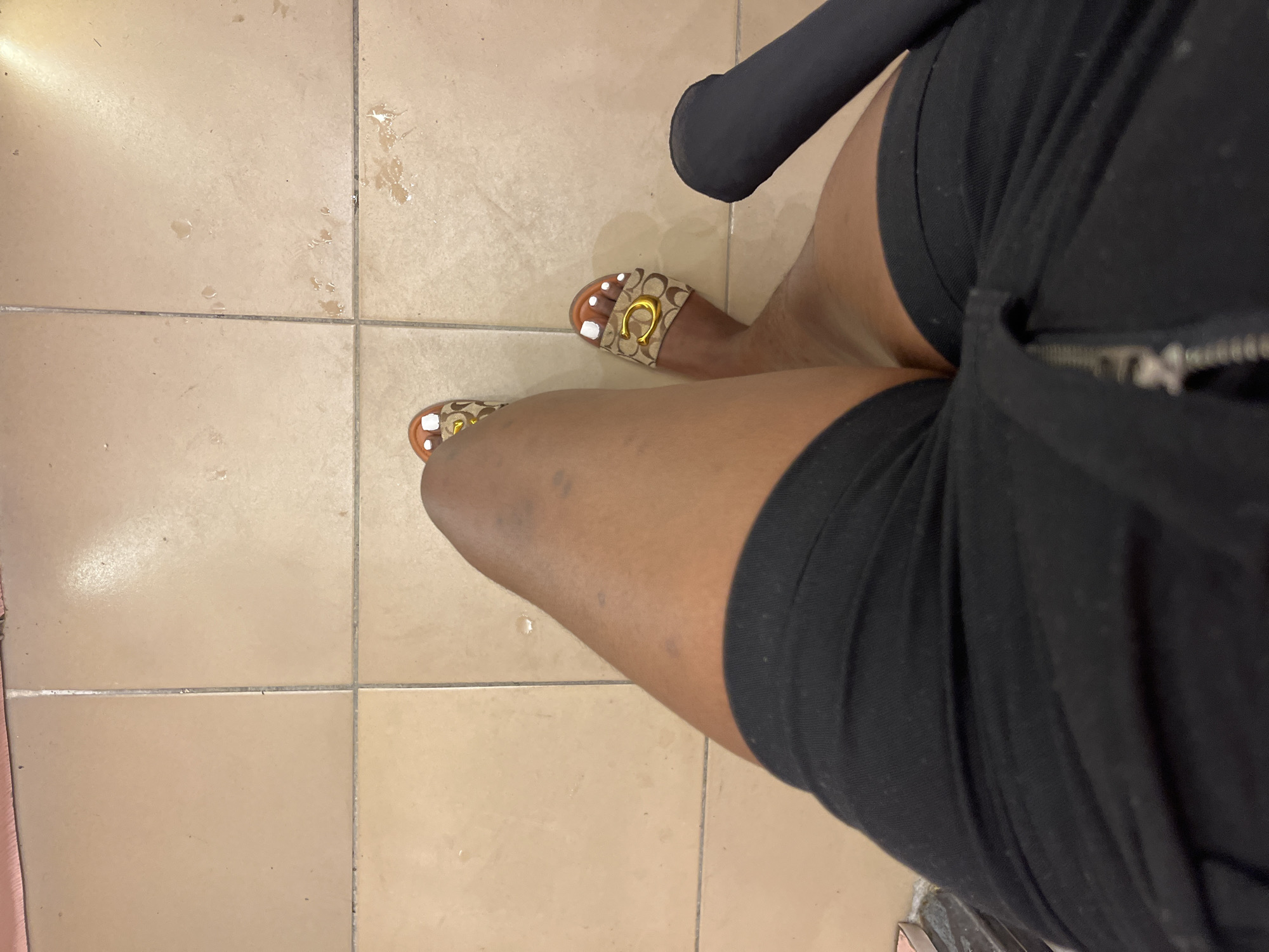 The image shows a close-up view of a person's legs and feet, wearing black shorts and slides with a distinctive "C" logo. The person is standing on a tiled floor, which appears to be in a domestic setting. The lighting suggests it might be daytime, and there are some water marks on the tiles, hinting at recent activity. The overall impression is casual and relaxed, with no specific event being depicted. The focus is on the fashion items and the textures of the clothing and the floor. The provided location context of Howayi, Nigeria, implies a warm climate where such attire would be appropriate. No text is visible in the image.