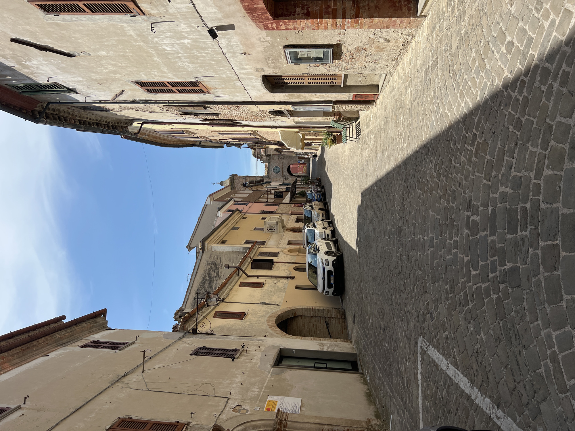 A cobblestone street runs between two rows of aged buildings in Barbara, Italy. The buildings on the left are primarily beige, with arched doorways and wooden shutters on the windows. On the right, the buildings are a mix of beige and brick, also featuring shutters and visible architectural details like exposed brickwork and arched entrances. Several cars are parked along the street, and a lone figure walks in the distance towards an archway that frames a clock tower. The sky above is clear and blue, casting distinct shadows across the pavement. A green bench sits outside one of the buildings on the right.