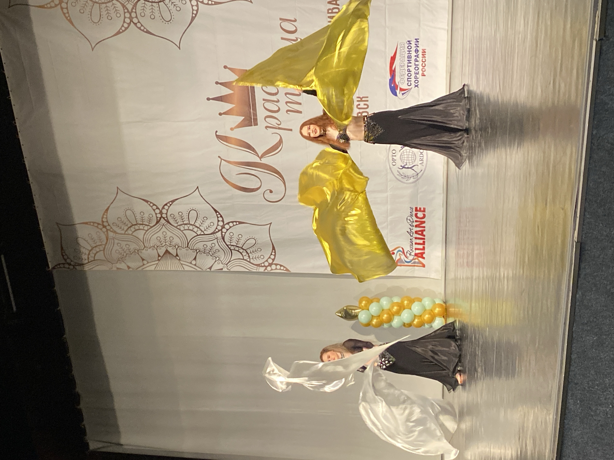The image captures a stage performance, likely a dance competition, based on the banner visible in the background. Two women are on stage, dressed in belly dancing attire. The first woman is located toward the top of the frame and wears a dark, embellished costume. She is holding two large, shimmering gold scarves in each hand and is captured in mid-motion. Below her, the second woman is in a similar dark costume, dancing with a long, white scarf. Behind the dancers is a white backdrop bearing designs, text, and logos including "Федерация Спортивной Хореографии России" (Federation of Sports Choreography of Russia), and "Russian Art Dance ALLIANCE." A string of balloons is placed on the stage near the second dancer. Given the location "Izhevsk, Russia" from the image's metadata, the scene is potentially occurring within that city.