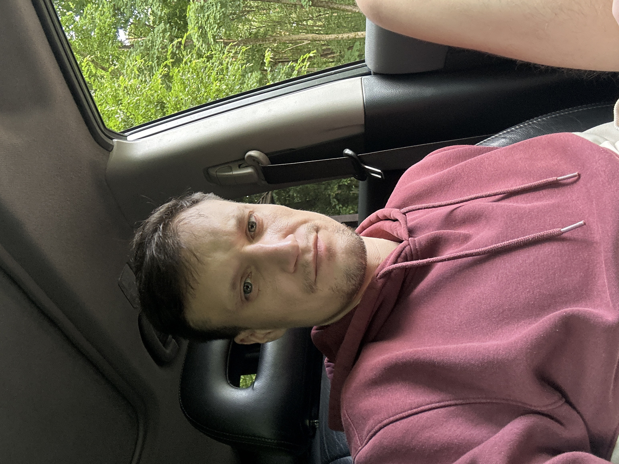 The image shows a man sitting in the driver's seat of a car, with the passenger window to his right. The man has light skin, short brown hair, and is wearing a maroon hooded sweatshirt. He is looking directly at the camera with a neutral expression. The interior of the car is visible, including the headrest of the seat behind him and the seatbelt buckle. Outside the window, there is lush green foliage and trees, suggesting a natural or rural setting. The lighting suggests it is daytime. The image does not provide any specific information about the location within the city of Shatava, Ukraine.