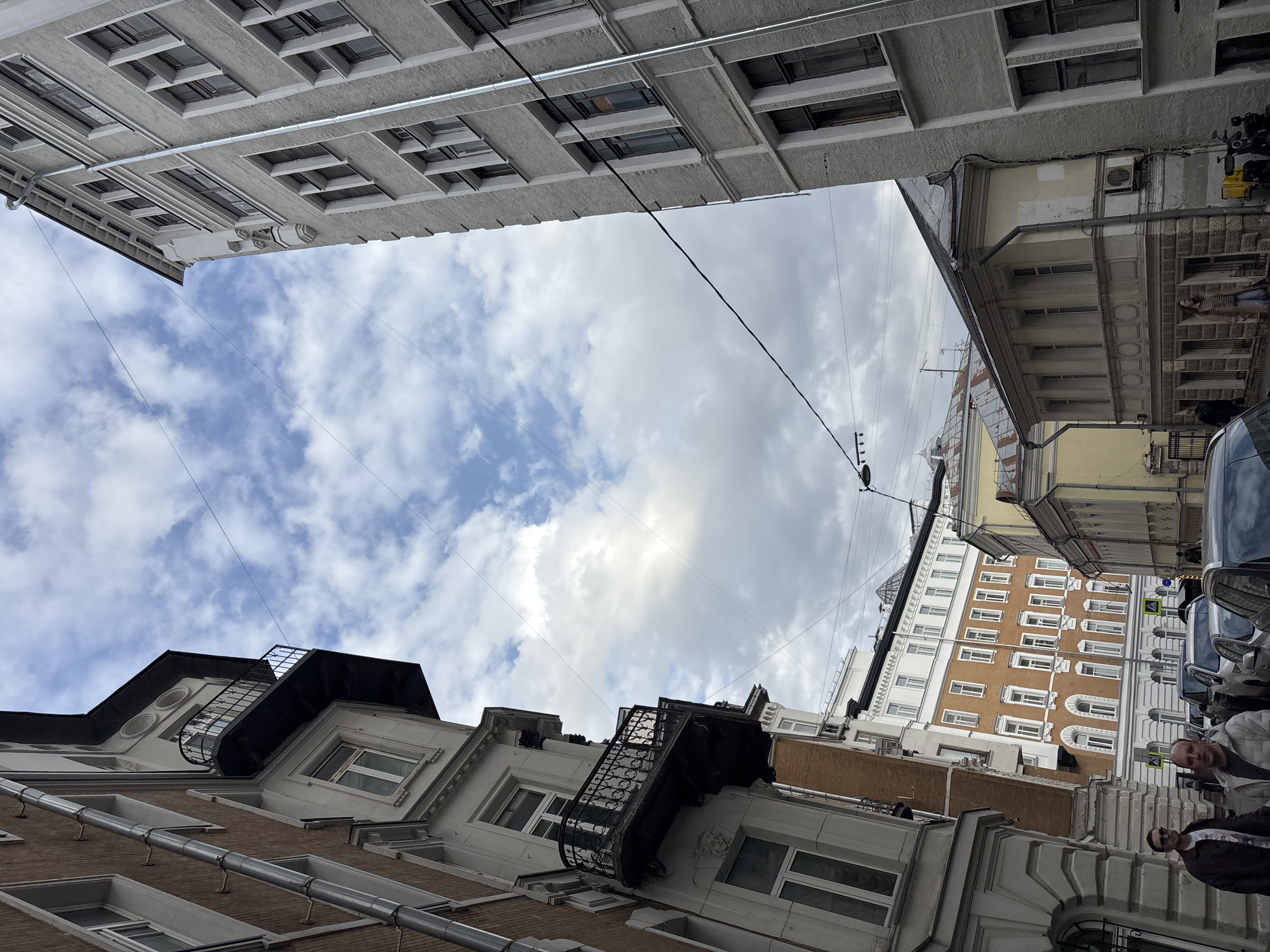 The image is taken from a low-angle perspective, looking up at the sky framed by buildings in what appears to be an alleyway. The sky is partly cloudy with patches of blue visible. The buildings exhibit a variety of architectural styles and colors, ranging from gray to beige and brown. Some buildings have detailed window frames and balconies. Power lines and other utilities crisscross the space between the buildings. On the right side of the image, people are visible, partially obscured by parked cars. This scene is located in the city of Moscow, Russia.