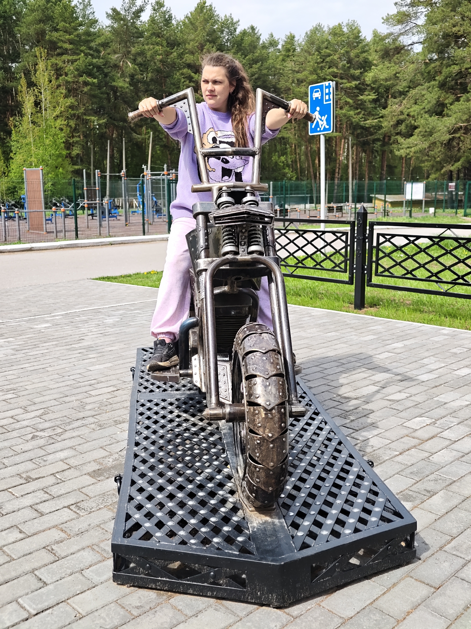 A young woman is sitting on a metal sculpture of a motorcycle on a black metal grid ramp in Chaikovsky, Russia. She wears a lavender sweatshirt with a cartoon graphic, pink sweatpants, and black sneakers. She is holding the handlebars, looking to the right of the frame. Behind her is a green fence, a green lawn, and a sign indicating a pedestrian crossing. In the background, trees and playground equipment can be seen. The area is paved with grey bricks.