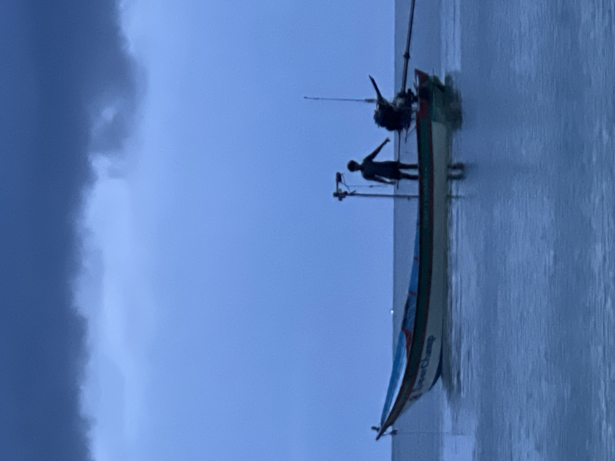 A silhouette of a lone individual stands in a small boat on a body of water under a stormy sky. The boat, which appears to be a traditional fishing vessel, is painted with green, white, and red stripes along its hull, and the word "Champ" is visible on its side. A small outboard motor is attached to the stern of the boat, and a fishing pole extends from the right side. The sky is dominated by dark, heavy clouds, suggesting an impending storm, with a lighter blue hue visible at the horizon. The water is calm, reflecting the muted colors of the sky and boat. The scene evokes a sense of solitude and anticipation, possibly capturing a moment before or during a fishing expedition amidst challenging weather conditions. This scene is likely located off the coast of Ko Pha-ngan District, Thailand, a region known for its picturesque beaches and fishing communities.