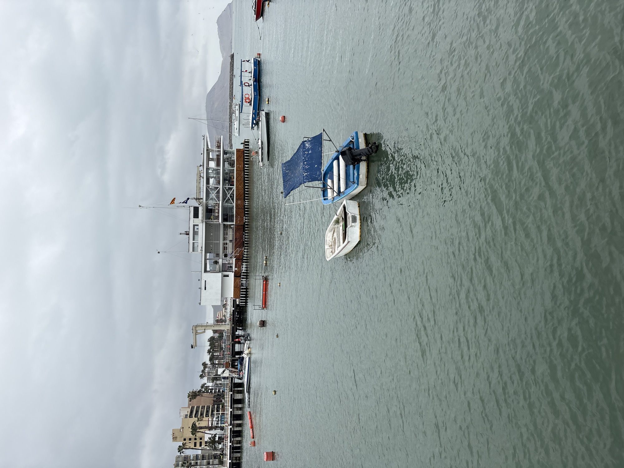 The image shows a cloudy day at a harbor in La Punta, Peru. In the foreground, two small boats are floating on the water. The boat on the left is white and appears to be unoccupied. The boat on the right is blue and white, with a blue canopy and an outboard motor. It also appears to be unoccupied.
In the midground, a larger white building with multiple levels and large windows is situated on a pier. A white sailboat is docked near this building. Several smaller boats and buoys are visible in the water around the pier.
In the background, several multi-story buildings with palm trees in front of them line the shore. Further in the distance, a mountain range can be seen under the overcast sky. Seagulls are flying in the sky. The water appears calm with gentle ripples.