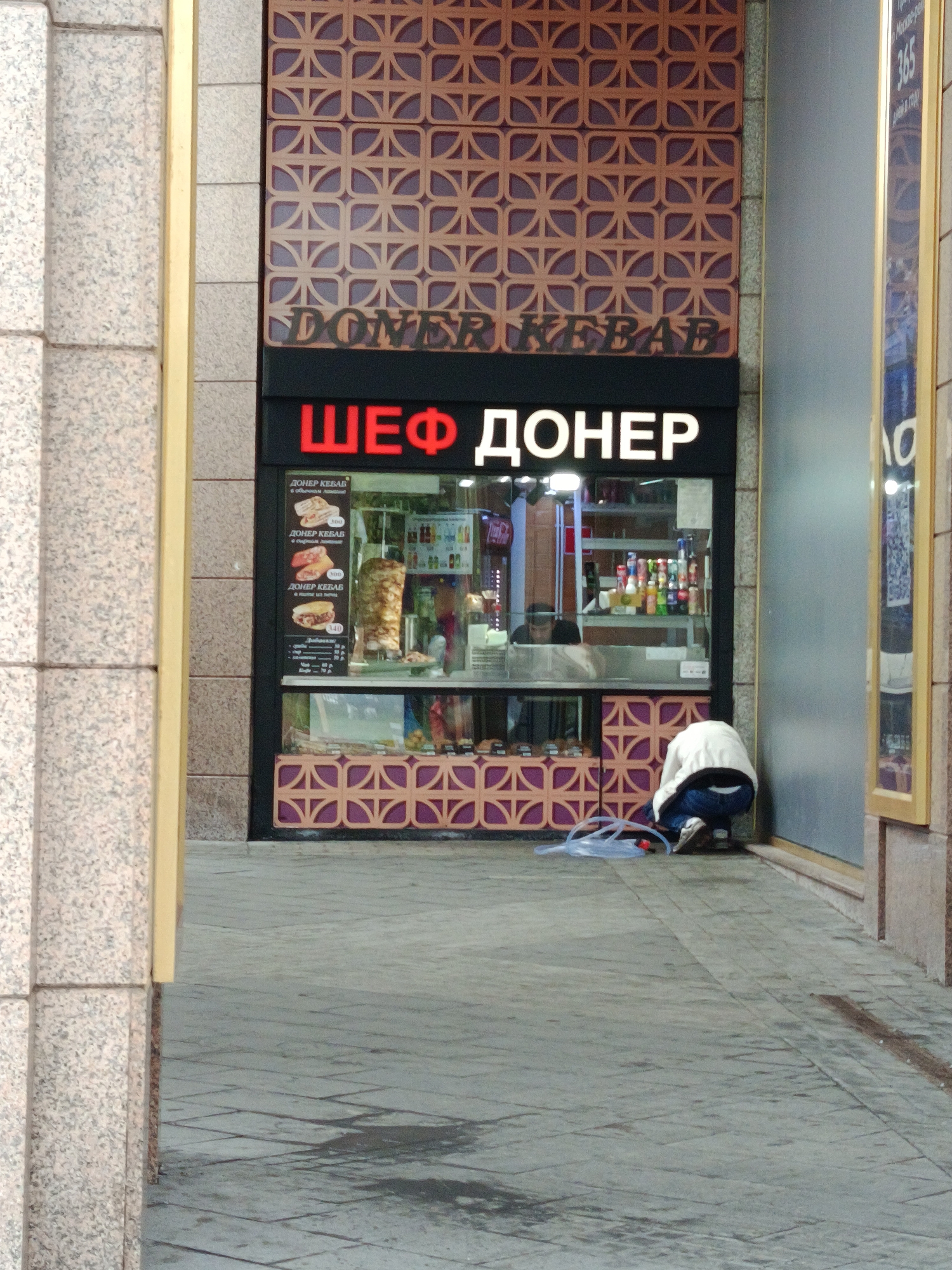 A street scene in Moscow, Russia, features a "ШЕФ ДОНЕР" (Chef Doner) kebab shop. The storefront has a sign in both English and Russian, displaying the menu and prices. A person is crouched down in front of the shop, near a blue object on the ground. The building facade has a patterned design above the shop and is flanked by columns on either side. The pavement in front of the shop shows signs of being damp.