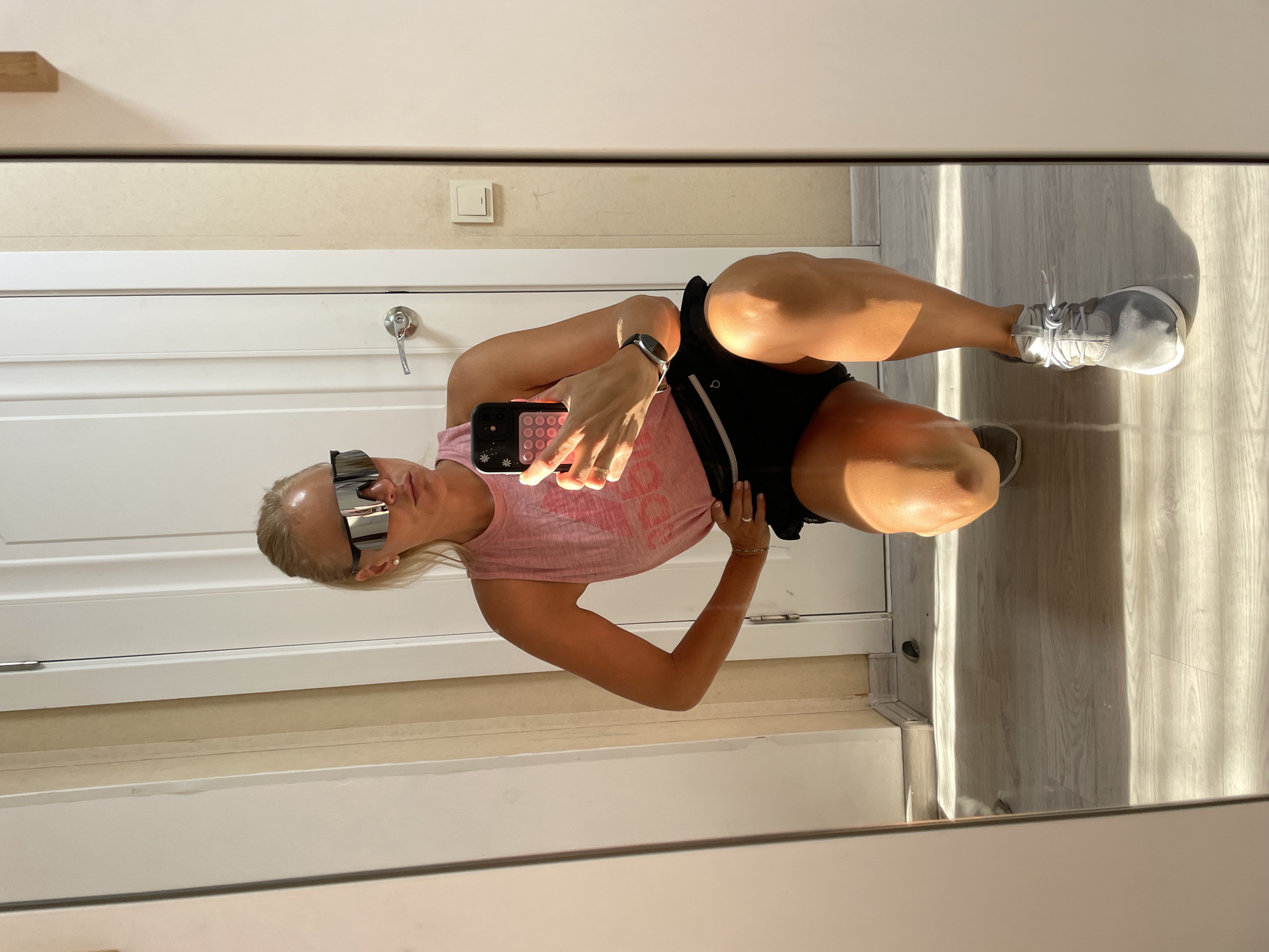 A woman wearing a pink tank top, black shorts, and sunglasses is posing for a mirror selfie. She is kneeling with one leg bent and the other extended. The room appears to be a hallway or entryway, with white doors and a light-colored floor. Sunlight is streaming in from the left, casting shadows on the floor and the woman's legs. The woman is holding a smartphone in her right hand, taking the picture. There is a light switch on the wall behind her. The image is set in the city of Baranovka, Russia.