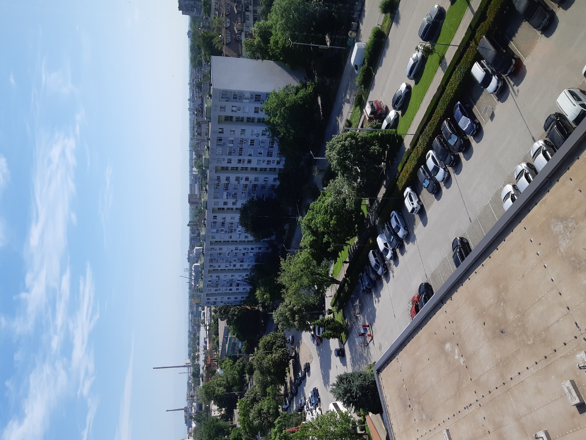 This image shows an elevated view of Plovdiv, Bulgaria. The foreground includes part of a flat rooftop, with a row of parked cars adjacent to it. Beyond the parking, the scene unfolds to reveal residential buildings, interspersed with green trees. Several chimneys are visible in the distance, suggesting industrial activity. The sky is a clear blue, lightly scattered with clouds.
