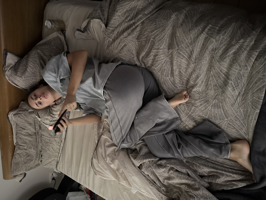 A single individual, appearing female, is observed lying on her back on a bed within an indoor residential setting. The specific location within the city of Warsaw, Poland, is not discernible from the visual evidence.
The individual, with dark hair pulled back, is clad in a light grey short-sleeved t-shirt and grey sweatpants. Her legs are bent and crossed, with her right foot resting on her left leg. Both feet are bare. She holds a black smartphone in her right hand, positioned towards her upper chest, with a bright pink reflection on its screen. Her left arm rests across her abdomen. Her gaze is directed towards the camera, with a neutral expression and slightly pursed lips.
The bed features a light-colored wooden headboard. The bedding consists of light beige sheets and two large pillows with a grey and white patterned cover, likely a leaf or fern motif, positioned against the headboard. A third pillow with the same pattern is beneath the individual's head. A matching patterned duvet partially covers the lower portion of her body and the right side of the bed. Another plain, light beige duvet or blanket is visible on the left side of the bed. To the right of the individual's head, resting on the patterned duvet, is a silver aluminum can, consistent with a Coca-Cola beverage can. A dark, indeterminate object is partially visible on the floor or furniture to the left of the bed. The lighting is consistent with an indoor environment.