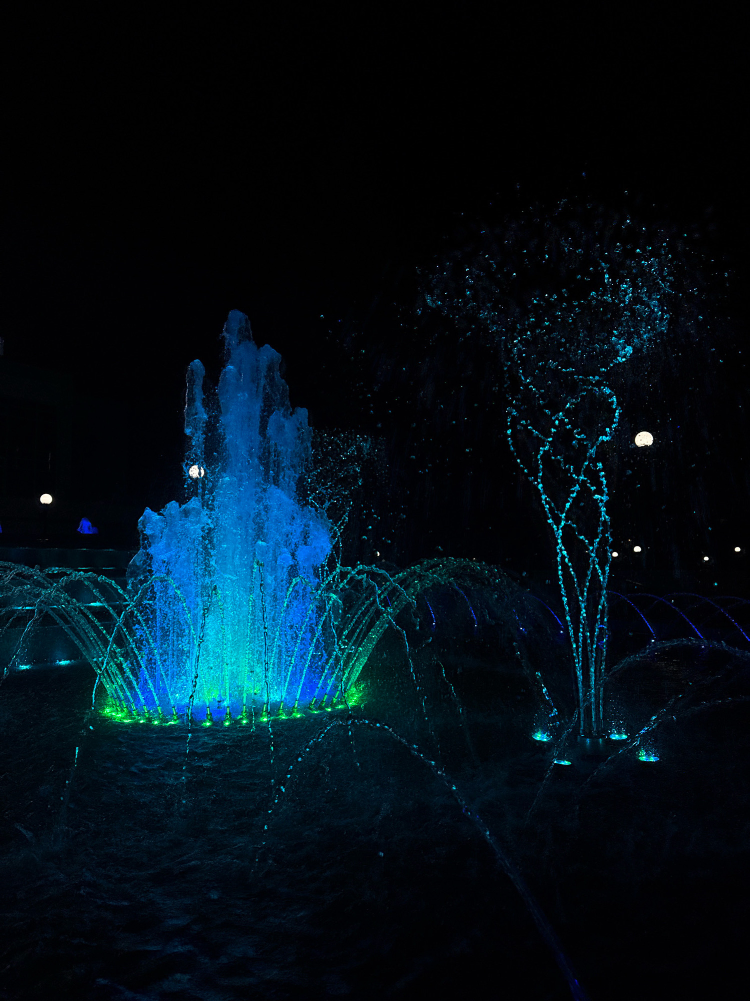 This is a nighttime photograph of a public fountain, likely in Mikashevichy, Belarus, illuminated by vibrant blue and green lights. The fountain features multiple water jets, with a large central column of water resembling a surge, flanked by smaller, arched streams and individual droplets of water creating a spray effect. The water is illuminated from below, casting a bright glow on the cascading liquid. The background is dark, suggesting it is nighttime, with a few distant, soft lights visible, possibly streetlights or architectural illumination. The overall impression is one of a dynamic and illuminated water display against a dark backdrop. There are no people or animals visible in the frame.