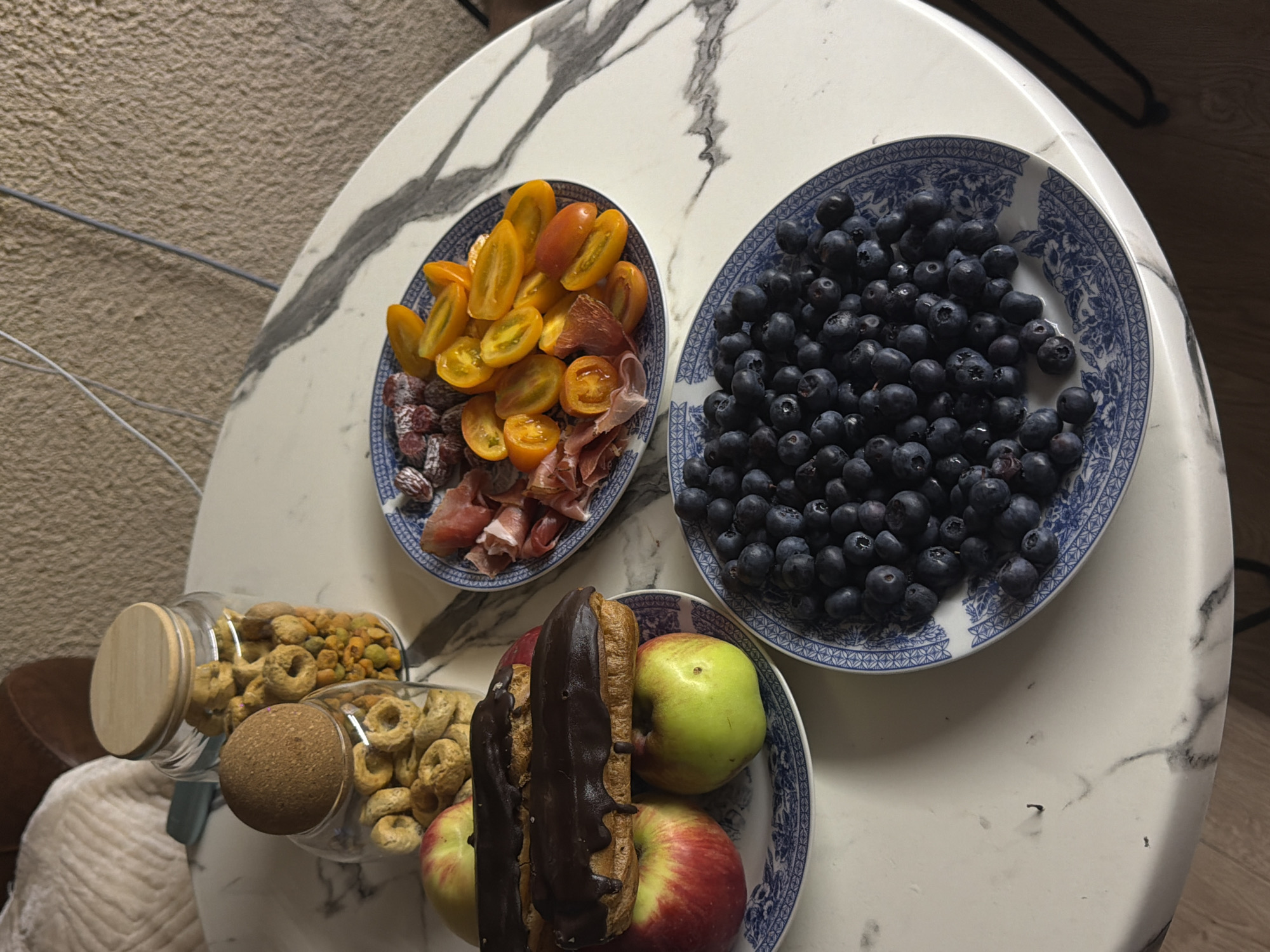The image displays a marble-top table set with various food items. On the table are two glass jars filled with circular, dry snacks. In front of the jars, on a plate with a blue and white pattern, are two chocolate-covered eclairs resting on two apples, one red and one green. To the right of the eclairs, on another plate with the same blue and white pattern, are sliced yellow cherry tomatoes, cured meat slices, and small sausages. A third plate with the blue and white pattern, positioned in the foreground, is filled with blueberries. The scene appears to be an indoor setting, possibly a home, with a textured wall in the background and a hint of wooden flooring visible at the bottom. There are no people or actions depicted in the image. The location in Tbilisi, Georgia, is not discernible from the visual information provided.