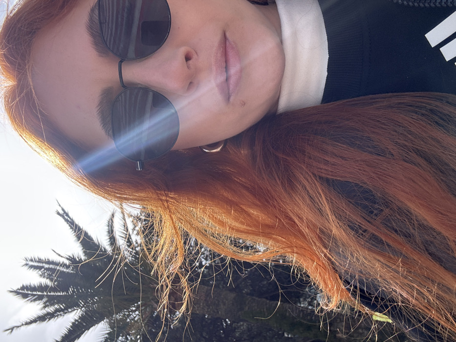 The image displays a close-up portrait of a female individual outdoors in Budva, Montenegro. The subject is presented from the mid-forehead to the lower chest, facing slightly to the right of the camera. She has long, reddish-orange hair, which appears wind-blown and is illuminated by direct sunlight, creating a prominent lens flare across the right side of the frame and reflecting off her dark, round-framed sunglasses. A small mole is visible on her upper lip area, inferior to her right nostril. She is wearing a light-colored turtleneck underneath a dark-colored top, which has a white logo partially visible on the upper right side. A gold-toned hoop earring is present on her visible left earlobe. In the upper left background, a large palm tree is silhouetted against a bright, possibly hazy, sky.