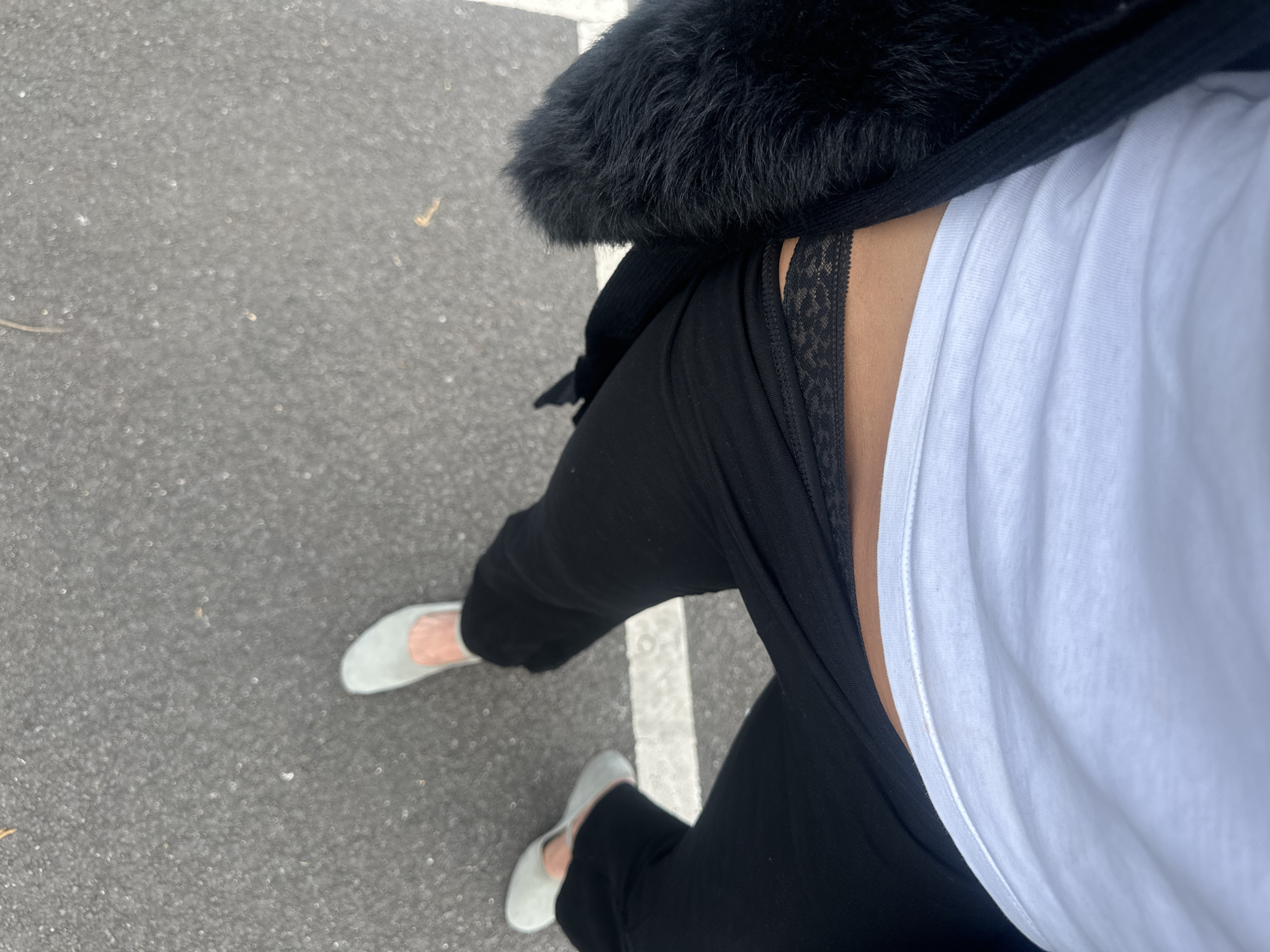 An aerial view depicts a person standing on asphalt, partially obscured by a white line. The individual is dressed in black pants and light-colored shoes. A white t-shirt is visible, along with a black jacket or coat featuring a fur collar. The location is a parking area or street in Tunbridge Wells, United Kingdom, based on potential contextual clues and visual evidence such as the style of the parking line and asphalt.