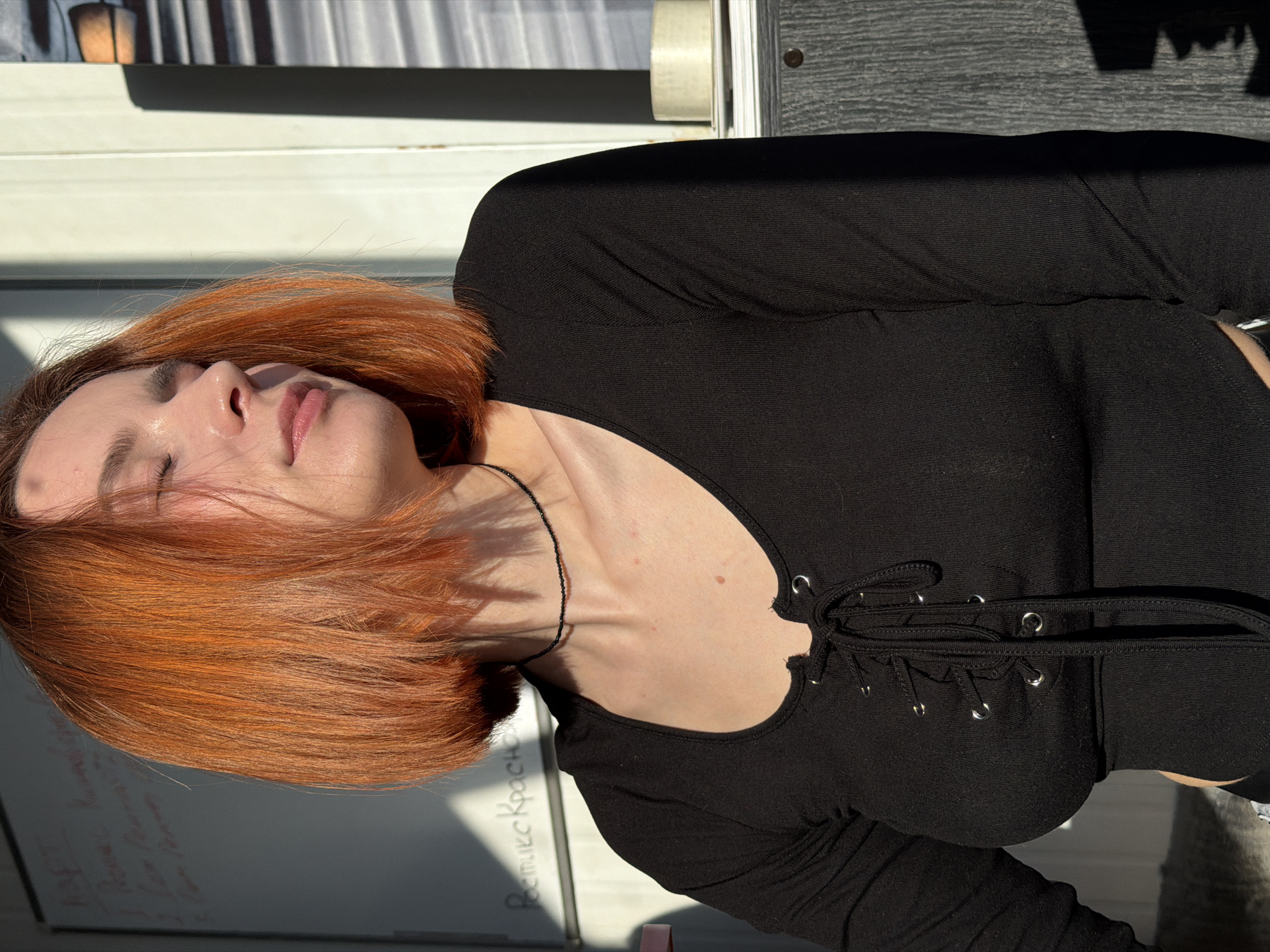 A woman with red hair, wearing a black top with a lace-up neckline, is reclining. She appears to be outside, with a window and possibly a table visible behind her. The location is Chaikovsky, Russia. There is handwriting in Russian visible on a surface near her head.
