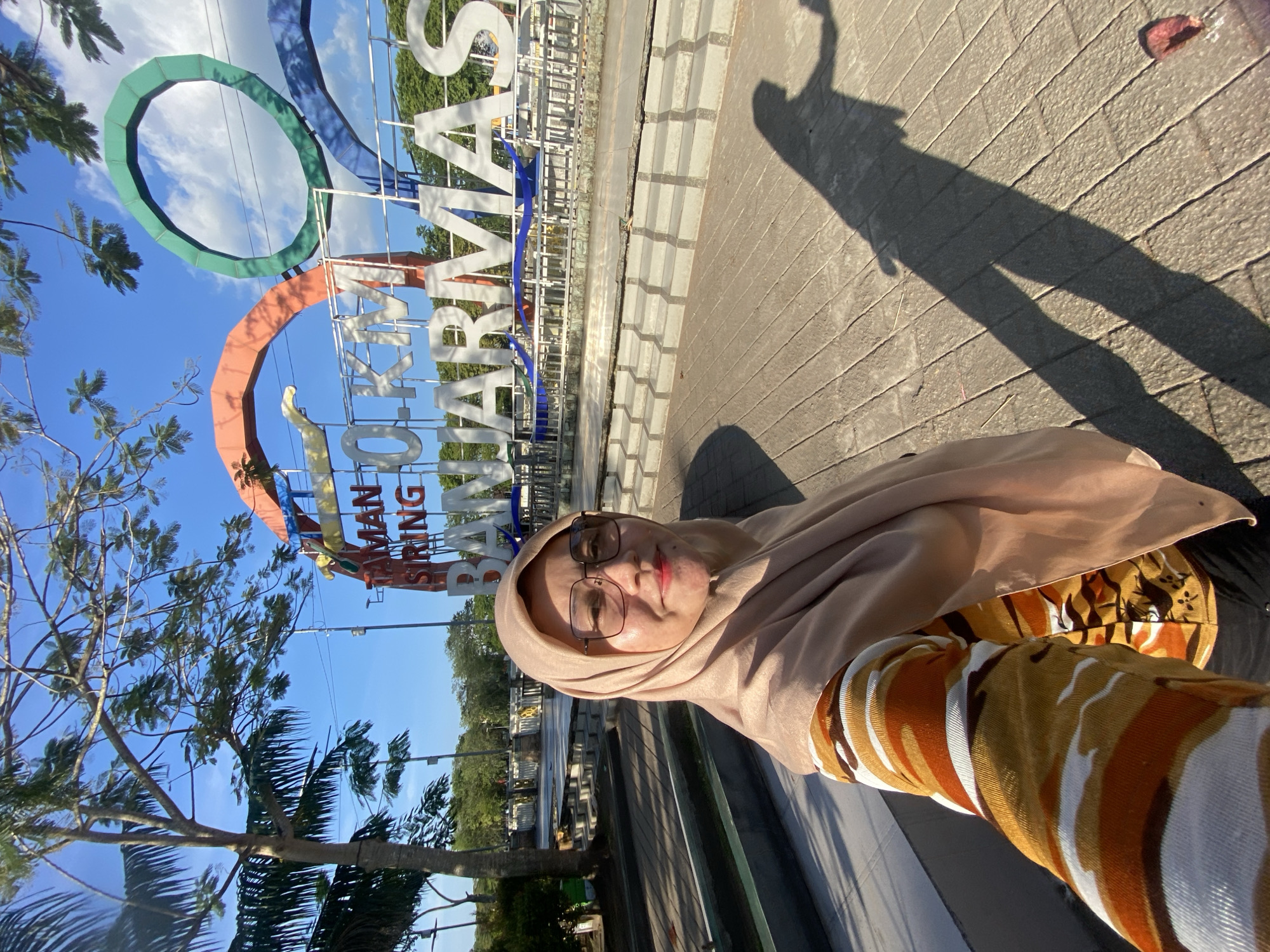 The image shows a woman taking a selfie in front of a large sign that reads "TAMAN 10 KM BANJARMASIN". The sign is colorful, with orange, green, and blue arches. The woman is wearing a beige hijab and glasses, and her shirt has a camouflage pattern in shades of orange, brown, and white. She is standing on a paved area with sunlight casting a long shadow of her onto the ground. In the background, there are trees, a clear blue sky with some clouds, and some stairs leading up to the sign. The scene appears to be a public park or recreational area in the city of Banjarmasin, Indonesia, likely commemorating a 10-kilometer park or pathway.