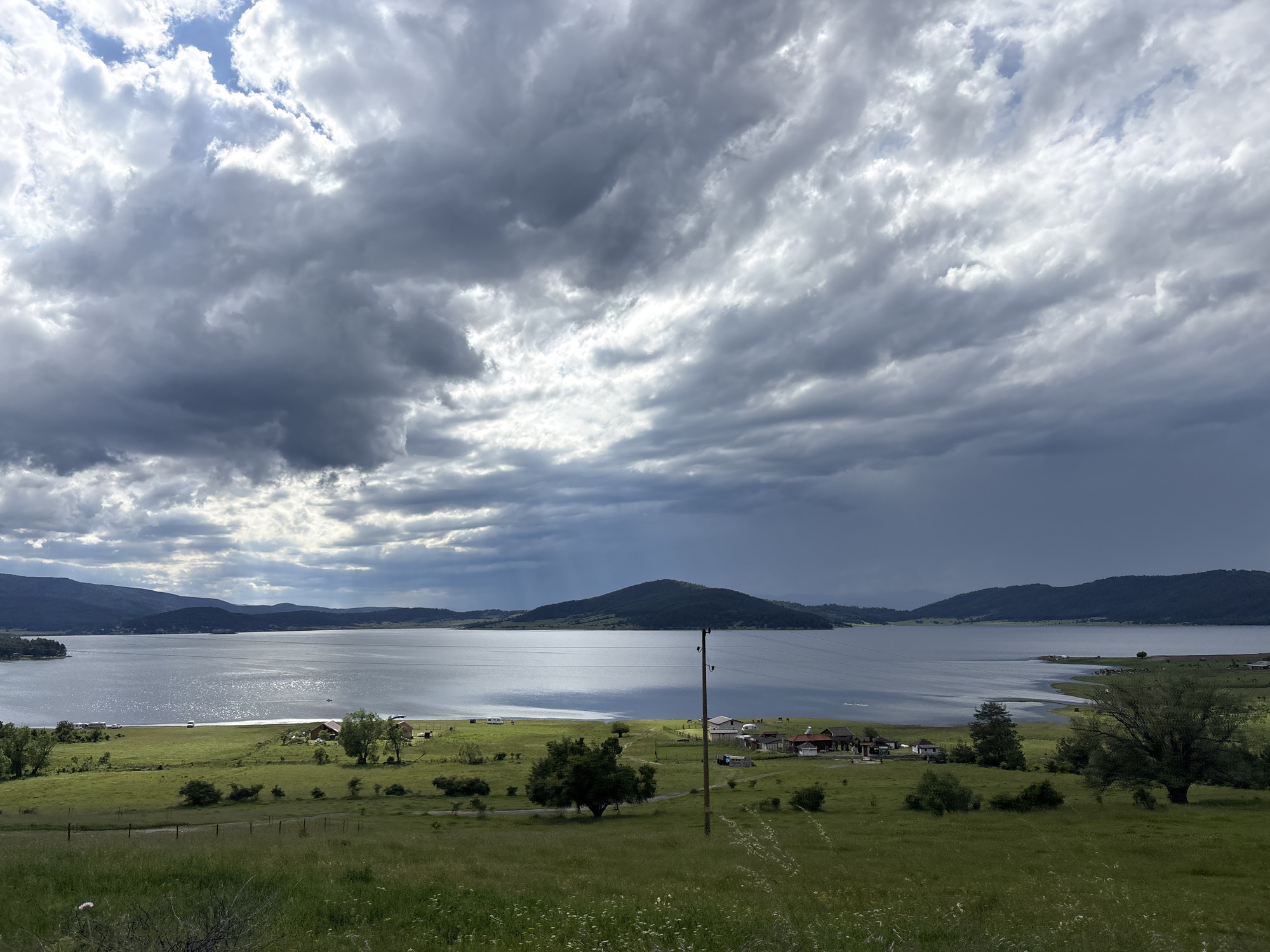 The image depicts a scenic view of the Batak Reservoir in Batak, Bulgaria. The reservoir occupies the central and background portions of the frame, its surface reflecting the dramatic, cloud-filled sky. The sky is characterized by a mix of dark, brooding storm clouds and patches of brighter, illuminated areas, with visible sunbeams or rain shafts descending towards the water.
The foreground and midground consist of rolling, verdant hills covered in green grass, dotted with wildflowers. A small cluster of buildings, likely a village or rural settlement, is visible along the shore of the reservoir. These buildings have red-tiled roofs and appear to be a mixture of houses and possibly other structures. There are a few scattered trees, some larger and more mature, and others smaller.
A wooden utility pole with wires runs vertically through the midground. In the water, near the shore, a few small boats or vessels are discernible. Further out on the water, the distant shores are lined with hills and mountains, their silhouettes softened by the atmospheric conditions. The overall impression is one of a natural landscape under a dynamic, changeable sky.