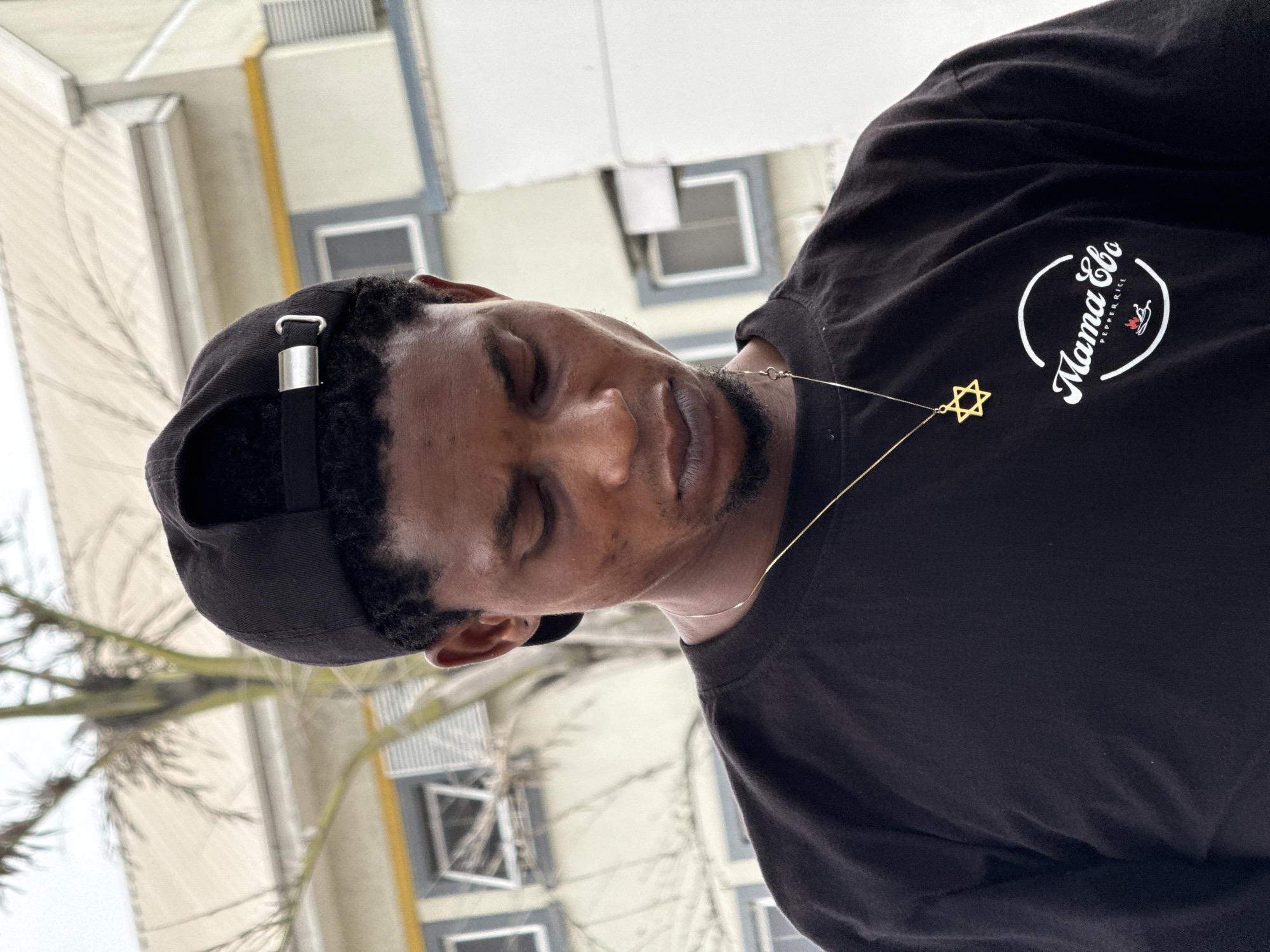 A man with dark skin and closed eyes wears a black t-shirt with the white words "Mama Ebo Pepper Rice" printed on it in a circular logo. He also wears a black baseball cap and a gold Star of David necklace. The background appears to be a multi-story building with windows. Tree branches are also visible. The location is likely in Lagos, Nigeria.