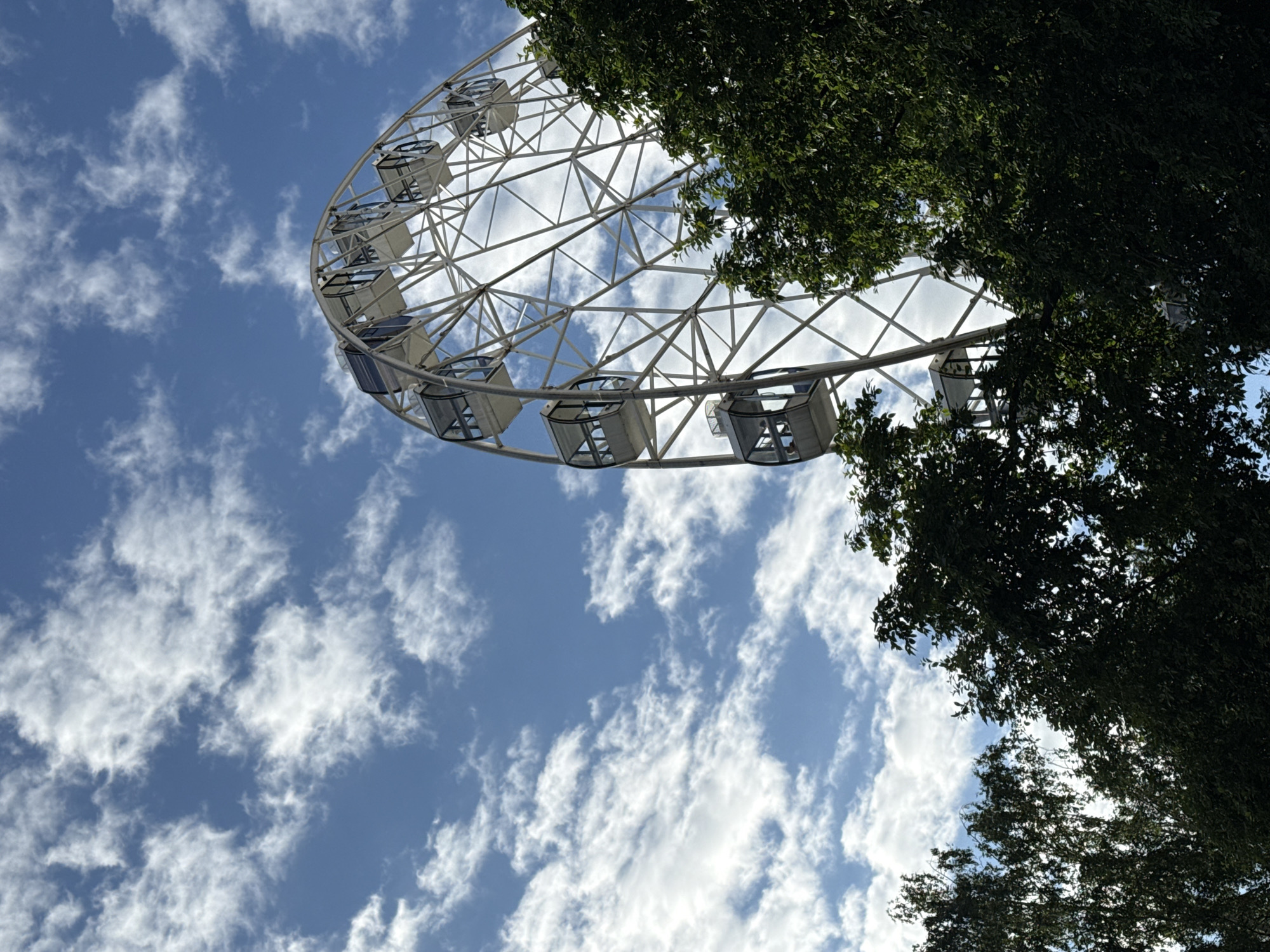 A Ferris wheel is visible in the upper right quadrant of the image, set against a bright blue sky with scattered white clouds. The Ferris wheel is predominantly white with a lattice-like structure and several enclosed cabins. Lush green trees with dense foliage occupy the lower portion of the frame, partially obscuring the base of the Ferris wheel. The image appears to be taken from a low angle, looking upwards. This Ferris wheel is likely located in the Summer Garden in Izhevsk, Russia, a popular recreational area.